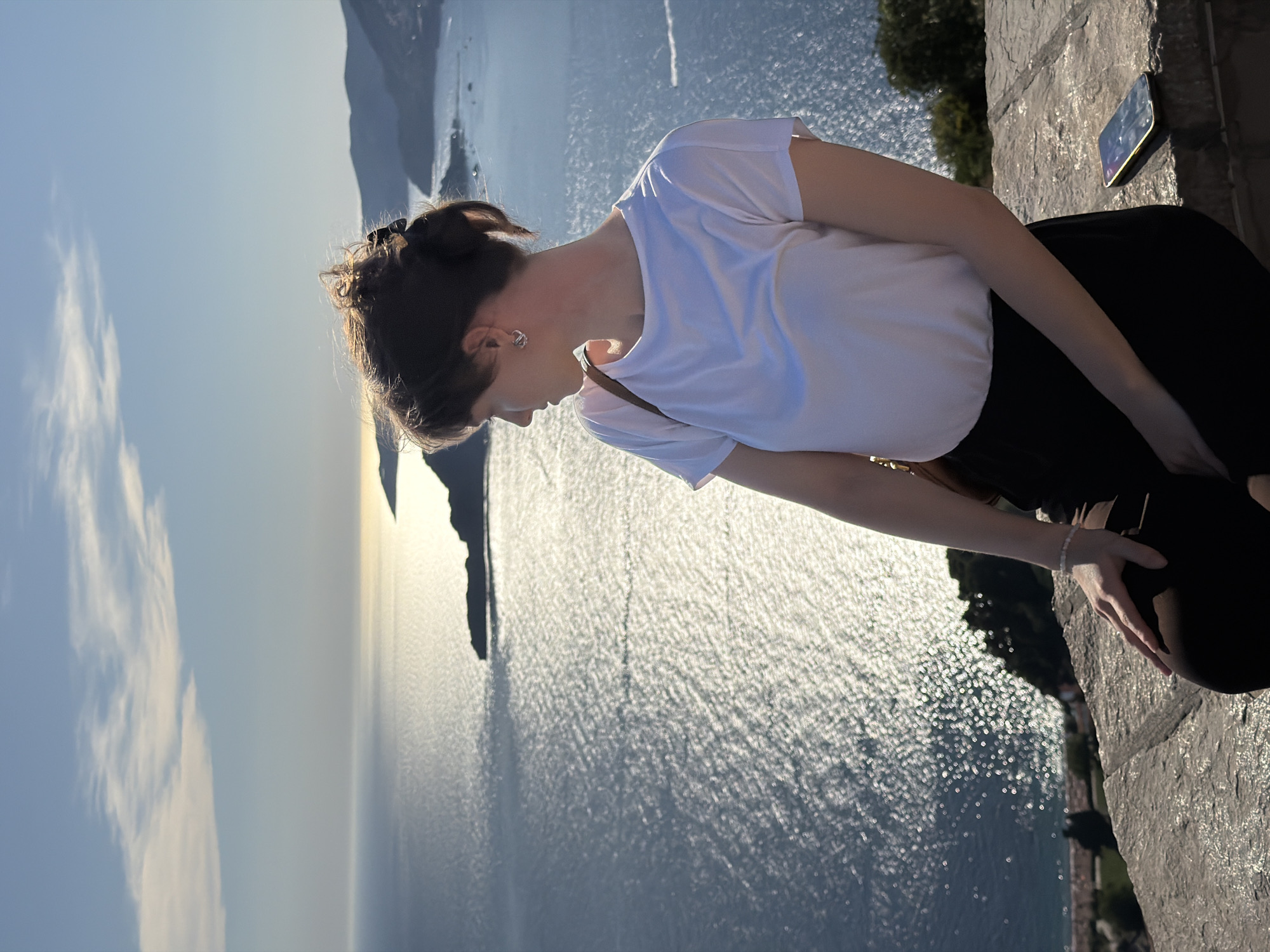 A woman with dark hair pulled back is seated on a stone ledge, looking out at a sun-drenched sea and coastline. She wears a white t-shirt and black pants. A brown bag strap is visible over her shoulder. A smartphone rests on the ledge to her right. The background features a body of water with shimmering sunlight, a small island with structures (likely Sveti Stefan), and distant hills. The sky is clear with a few wispy clouds. The overall scene suggests a serene, scenic view.