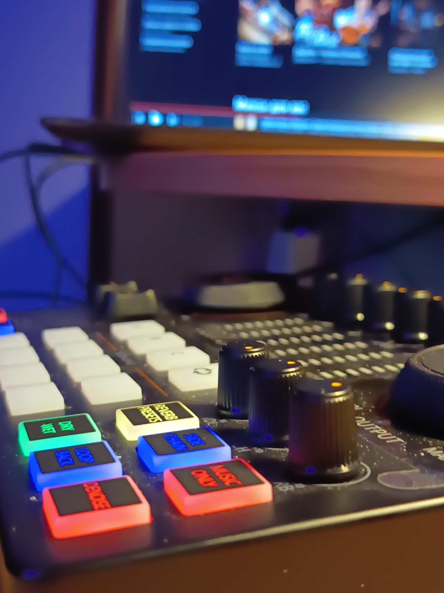 The image shows a close-up view of a DJ controller with various illuminated buttons and knobs. Some buttons are labeled with words like "DRY WET," "ZOOM BACK," "DECODE," "REVERB PRESETS," "SIDE CHAIN," and "MUSIC ONLY." The controller is positioned in front of a blurred background that includes a computer screen displaying what appears to be software with glowing blue elements. The lighting suggests a dim environment, with the DJ controller's lights being the primary light source in the foreground. There are no people visible in the image, and the scene appears to be indoors, likely in a home studio or similar setup. The location of Izmail, Ukraine, is not depicted or suggested by any elements within the image.