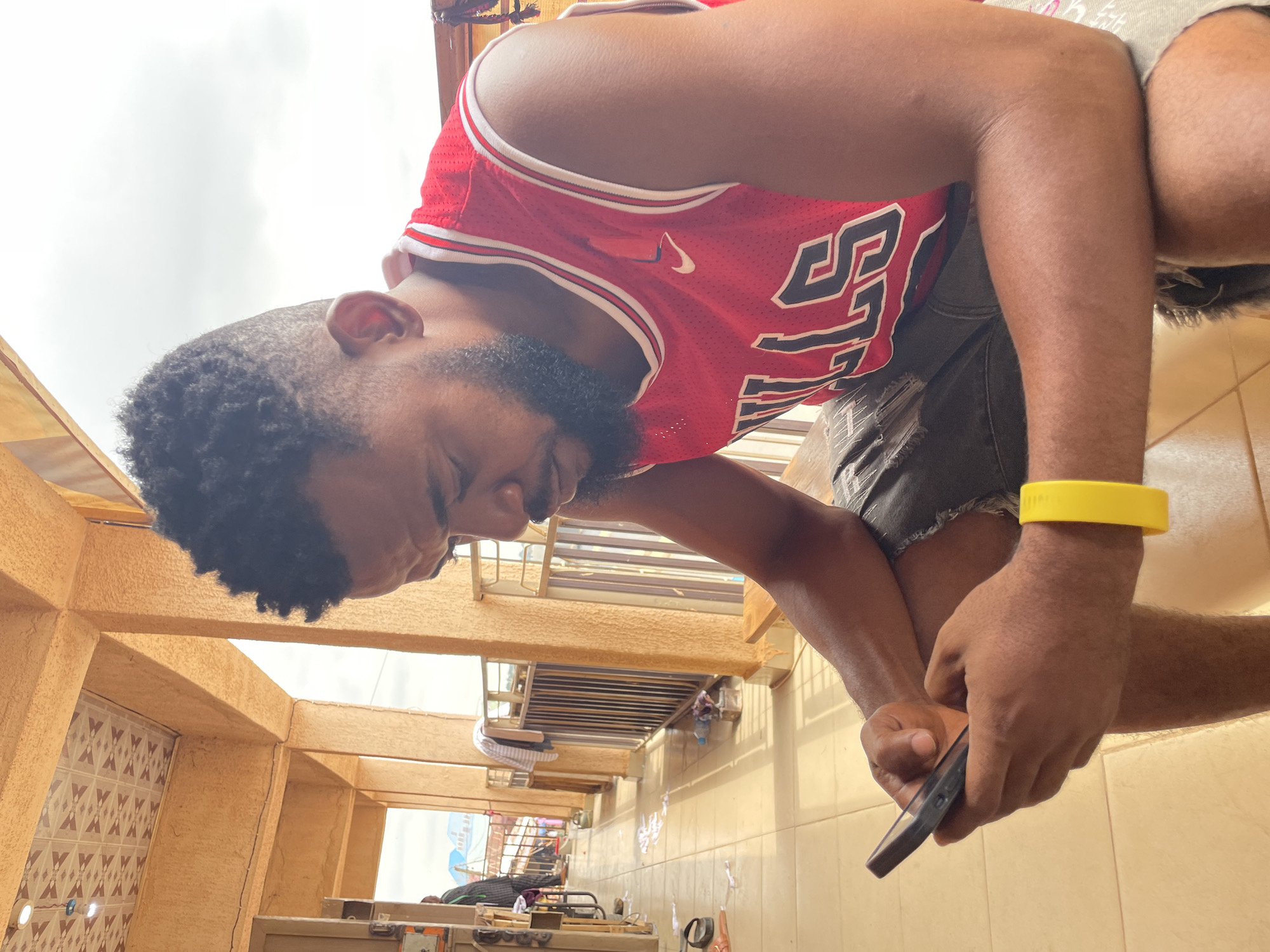 A medium shot depicts a dark-skinned man with a dark curly afro and a full beard, positioned on what appears to be a balcony or raised platform. He is wearing a red Chicago Bulls jersey with white trim and the number "23" on the front. The man is also wearing ripped grey shorts. He is looking down and holding a dark-colored cell phone in his hand. A yellow wristband is visible on his wrist.
The balcony is constructed of textured beige material and features a tiled floor. Through the balcony's opening, a partial view of what seems to be an outdoor market or street scene is visible. This is Akwanga, Nigeria, based on the location requested.