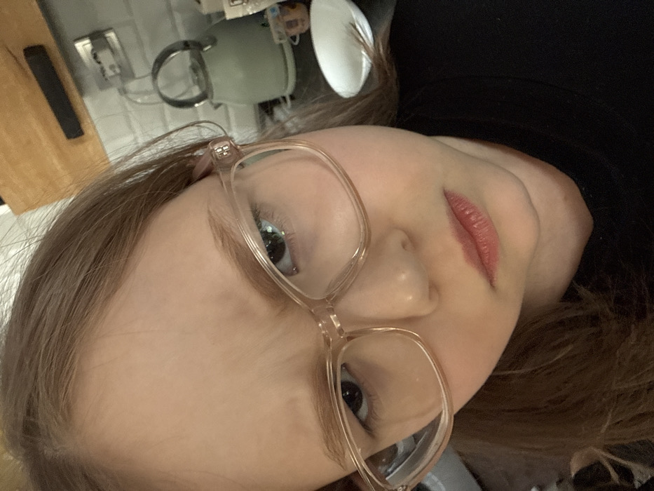 A close-up image shows a young female with light brown hair, wearing clear or light-colored framed eyeglasses. She looks directly at the camera with furrowed eyebrows and a downturned mouth, conveying a stern or displeased expression. The subject is wearing a dark-colored, likely black, shirt or garment. The background indicates an indoor setting, specifically a kitchen in the city of Кипчак, Russia. Visible elements on the right side of the frame include a light green or grey kettle with a metallic handle resting on a dark countertop, a white plate, and a tiled wall, possibly with subway tiles. Above the tiled section, a wooden shelf or cabinet is present, with a dark rectangular object mounted underneath it. A power outlet is also visible on the tiled wall.
