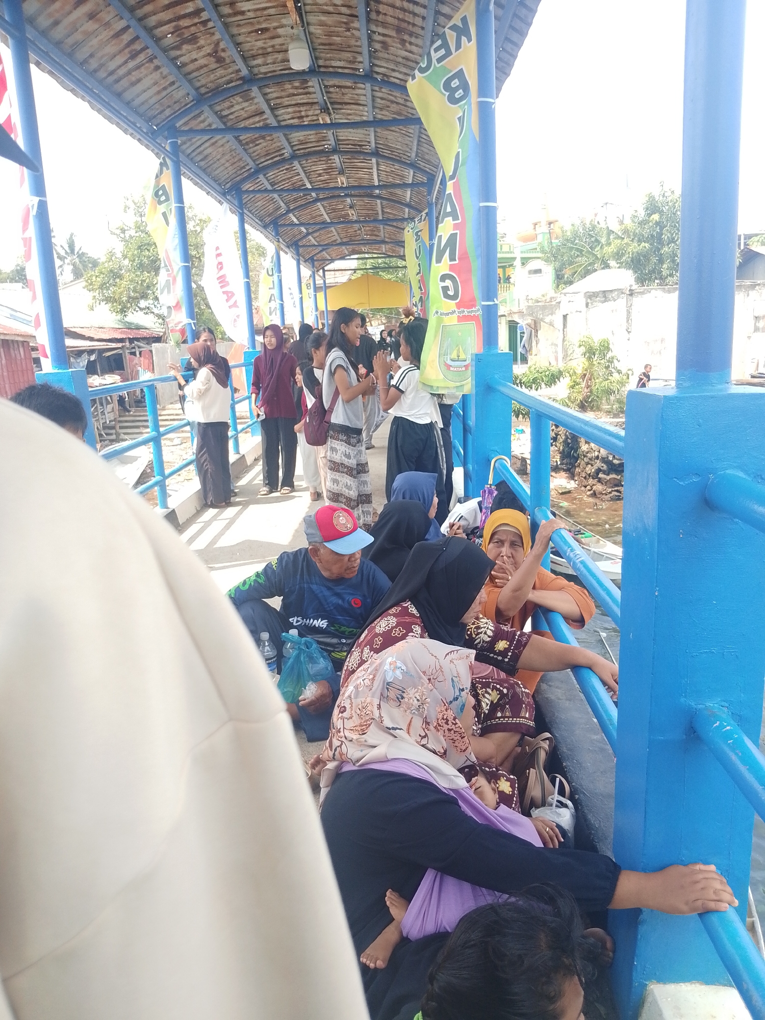 The image captures a bustling scene at a ferry terminal in Batam, Indonesia, likely near the waterfront. A group of people, predominantly women and some men, are gathered on a covered walkway or pier, waiting. The walkway features a blue metal railing and a corrugated metal roof supported by beams. Banners with text are visible hanging from the ceiling.
In the foreground, an elderly man wearing a blue shirt with "FISHING SPORT" written on it and a red and blue cap is seated on the ground, looking to his right. Next to him, a woman in a patterned headscarf and dark clothing is also seated, holding onto the blue railing. Another woman in a yellow headscarf is leaning on the railing, looking towards the water. Several other women in headscarves and varying attire are seated or standing, some appearing to be conversing or watching. In the background, a few younger women and girls are standing, some in casual clothing. Children are also present, one sleeping in a purple cloth. The environment suggests a warm, possibly humid day, with some greenery visible in the distance beyond the structures. The water is visible beyond the railing, with a small boat docked nearby. The overall impression is one of transit and waiting at a local transportation hub.