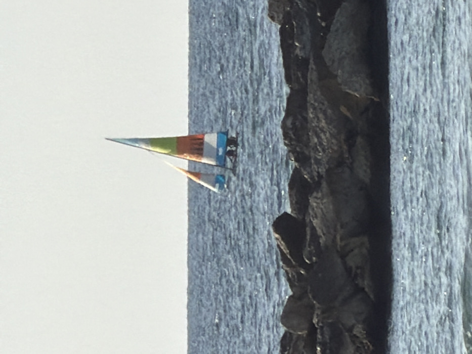 The image depicts a daytime scene on the Mediterranean Sea off the coast of Tel-Aviv, Israel. In the foreground, a dark rock breakwater spans horizontally across the lower portion of the frame, partially submerged in rippled blue-grey water. Beyond the breakwater, the open sea extends to a distant, flat horizon line, with moderate surface chop and sunlight reflections. In the midground, a small catamaran sailboat is underway. The catamaran features a main sail with horizontal stripes of light blue, white, orange, and lime green, and a smaller, triangular jib sail primarily orange and light blue. Two indistinct, dark figures are visible seated or crouched on the boat's hull, positioned beneath the main sail, operating the vessel. The sky above the horizon is clear and light-toned, indicating bright daylight.