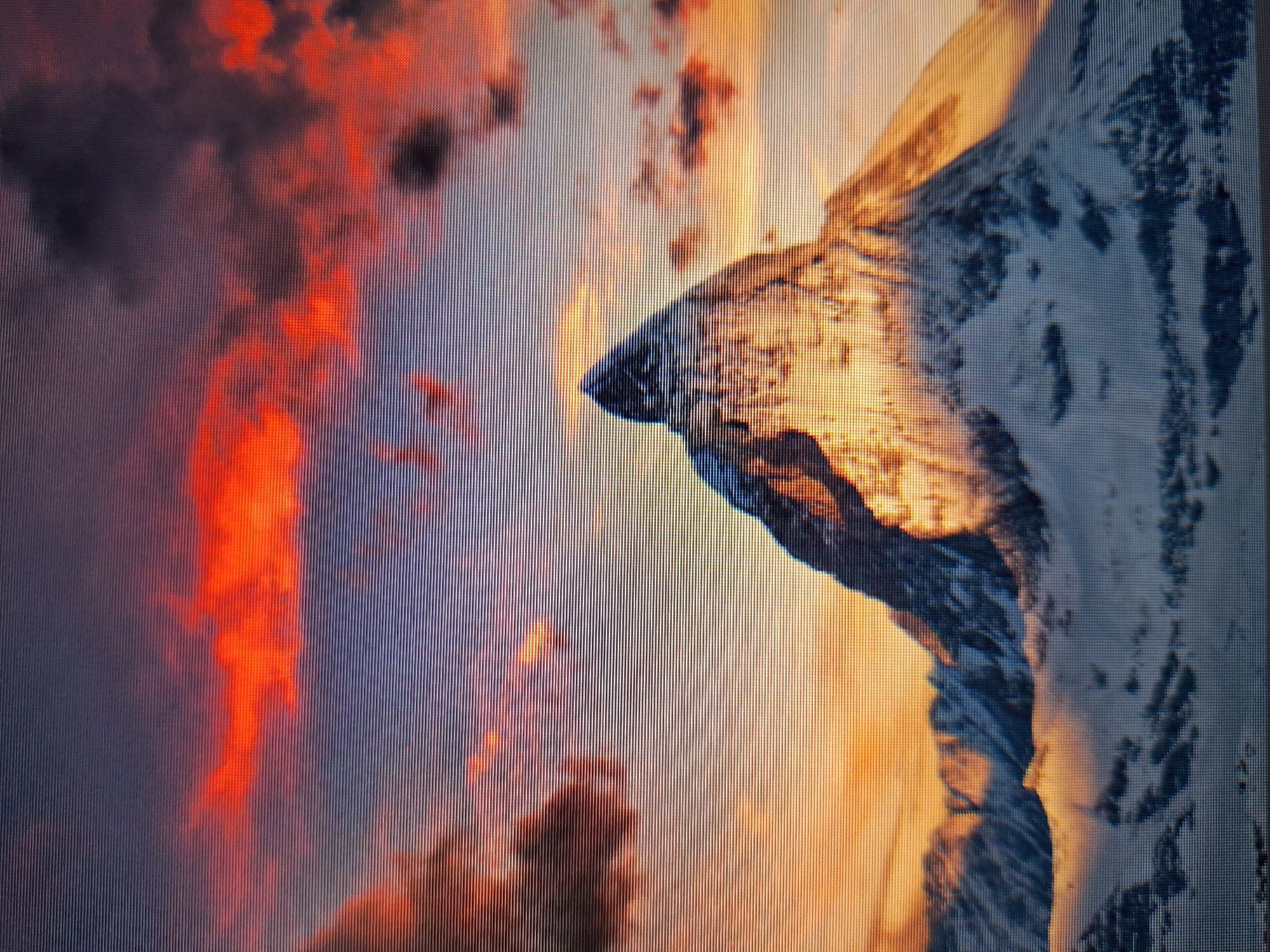 The image displays a majestic, snow-capped mountain peak set against a dramatic sunset sky. The mountain's summit is illuminated by the warm hues of the setting sun, casting a golden glow on its snowy slopes. The sky is a vibrant mix of fiery oranges, reds, and purples, with dark clouds adding depth and contrast. The foreground shows the lower slopes of the mountain, covered in snow and shadows. This image is not located in the city of Varna, Bulgaria.