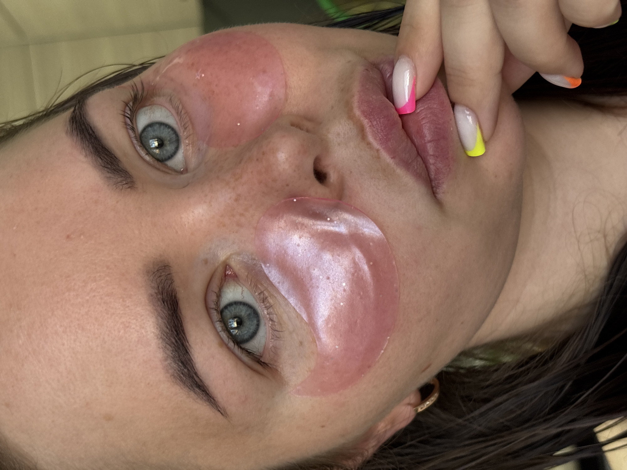 A close-up, high-angle portrait captures a young woman engaged in a skincare routine, likely indoors in Izhevsk, Russia. Her face is angled upwards and slightly to the side, with her striking blue-grey eyes looking towards the viewer. She is wearing two pink, shimmery hydrogel eye patches, positioned beneath each eye. Light freckles are visible across her nose and cheeks, and her eyebrows are dark and well-defined.
Her right hand is raised, with fingers gently touching her lips. A prominent detail is her vibrant, multi-colored French tip manicure, featuring tips in hot pink, neon yellow, and bright orange over a natural or clear base. A small, gold-colored hoop earring is visible in her right earlobe, and dark hair frames her face. The background is softly blurred, emphasizing the subject and the details of her self-care activity and colorful nail art. No text is visible in the image. The scene conveys a sense of relaxation and personal grooming.