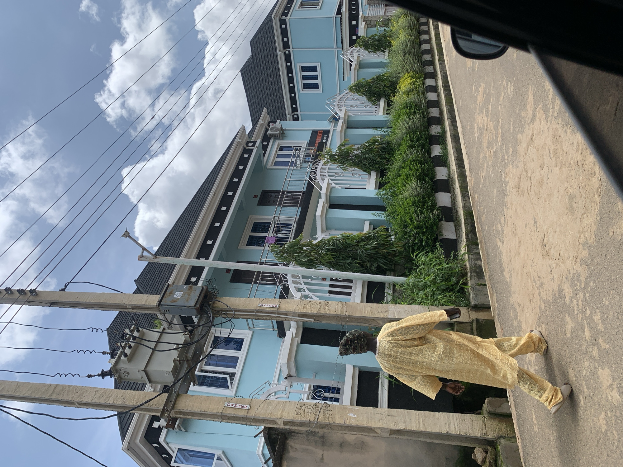 The image shows a street scene in an urban environment, possibly in Africa, given the attire of the individual. A man wearing a traditional yellow embroidered garment walks along an asphalt road. He is facing away from the camera and is positioned on the left side of the road. In the background, there are two modern, light blue buildings with dark roofs, featuring multiple windows and balconies. Electrical poles with a transformer and wires are prominent on the left side of the street, in front of the buildings. Lush green bushes and trees line the front of the buildings, separated from the road by a black and white curb. The sky is partly cloudy. The image appears to be taken from a moving vehicle, as indicated by the blurred black frame on the right side of the image.