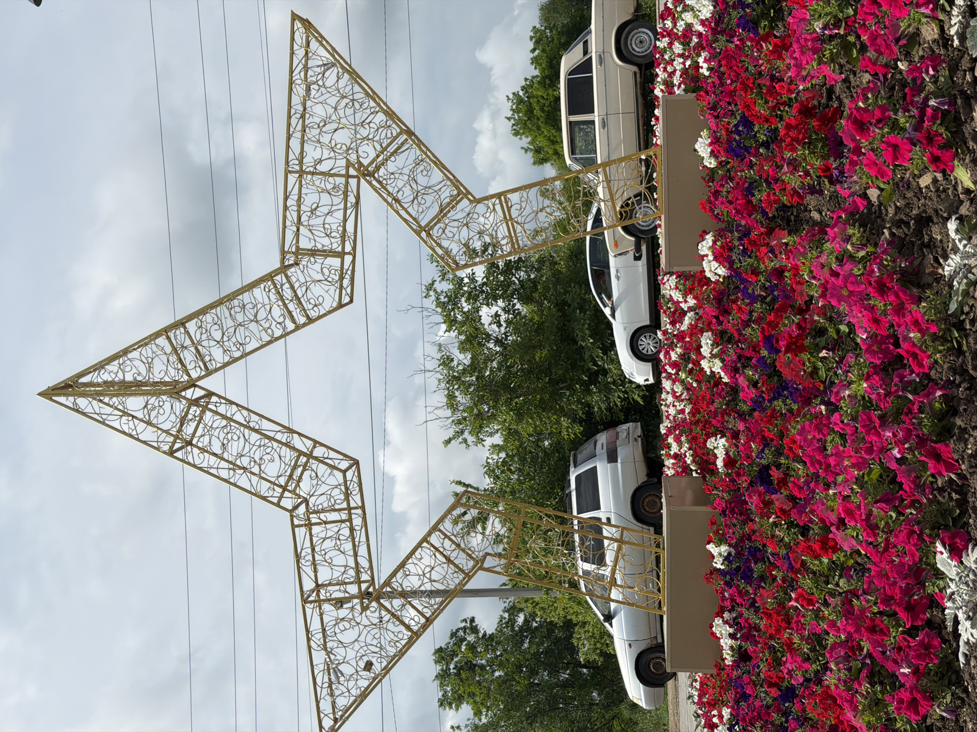 The image shows a large, golden, ornate, five-pointed star decoration in Leningradskaya, Russia. It's constructed from a metal framework with decorative scrollwork filling the interior spaces. The star is positioned upright and dominates the foreground. Behind the star, there are several cars parked near a flowerbed filled with red, white, and purple flowers. Green trees fill the middle ground behind the star and cars. Overhead, a cloudy sky is visible, with power lines stretching across the upper portion of the frame.