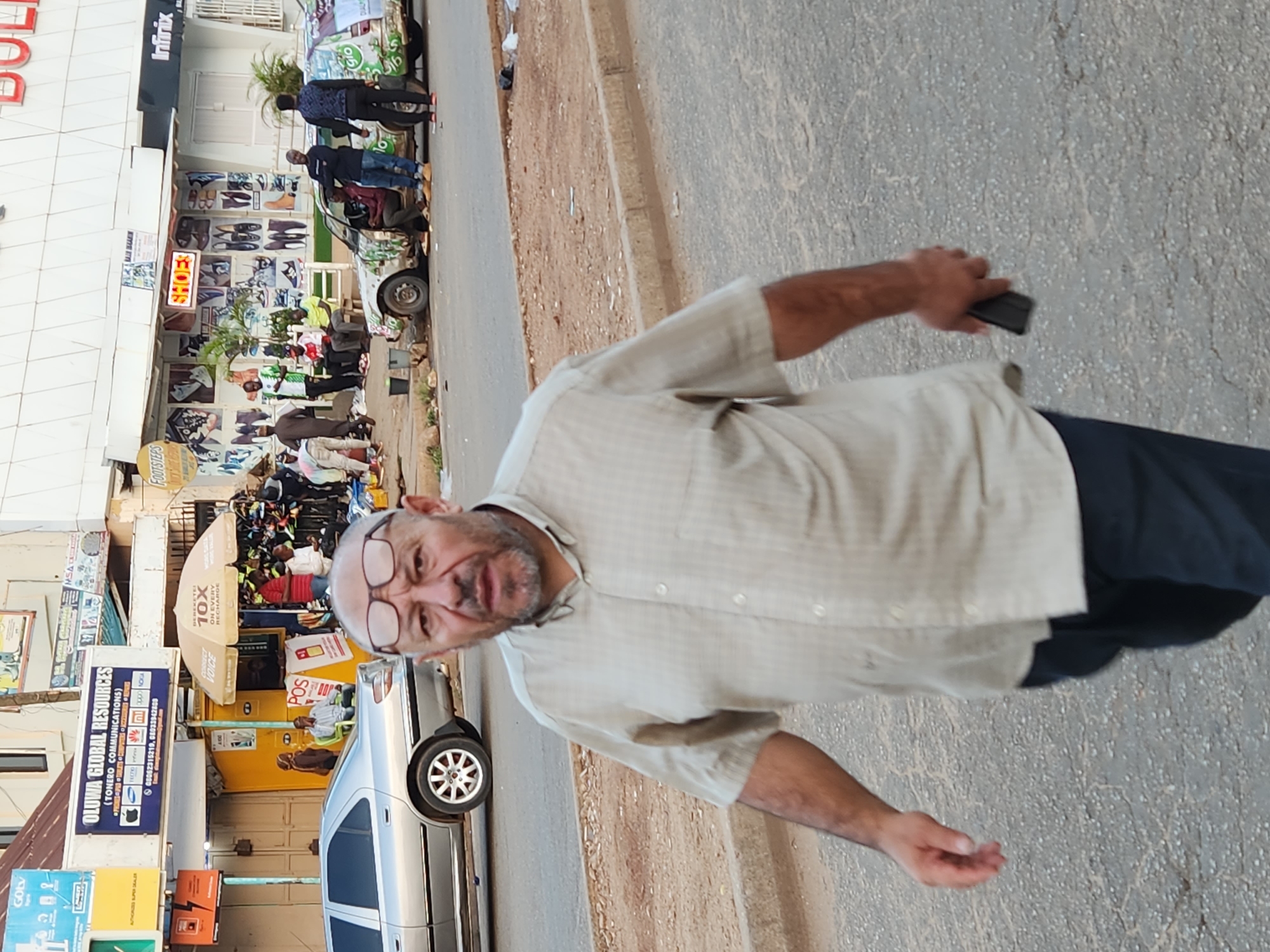 An eye-level portrait shot depicts a man with glasses and a tan shirt walking on a street. He appears to be holding a black object in his right hand. In the background, there are businesses, including a shoe store with images of shoes on display, and an umbrella stand. Cars are parked along the street, and pedestrians can be seen near the buildings. Various signs and advertisements are visible on the buildings and stands.