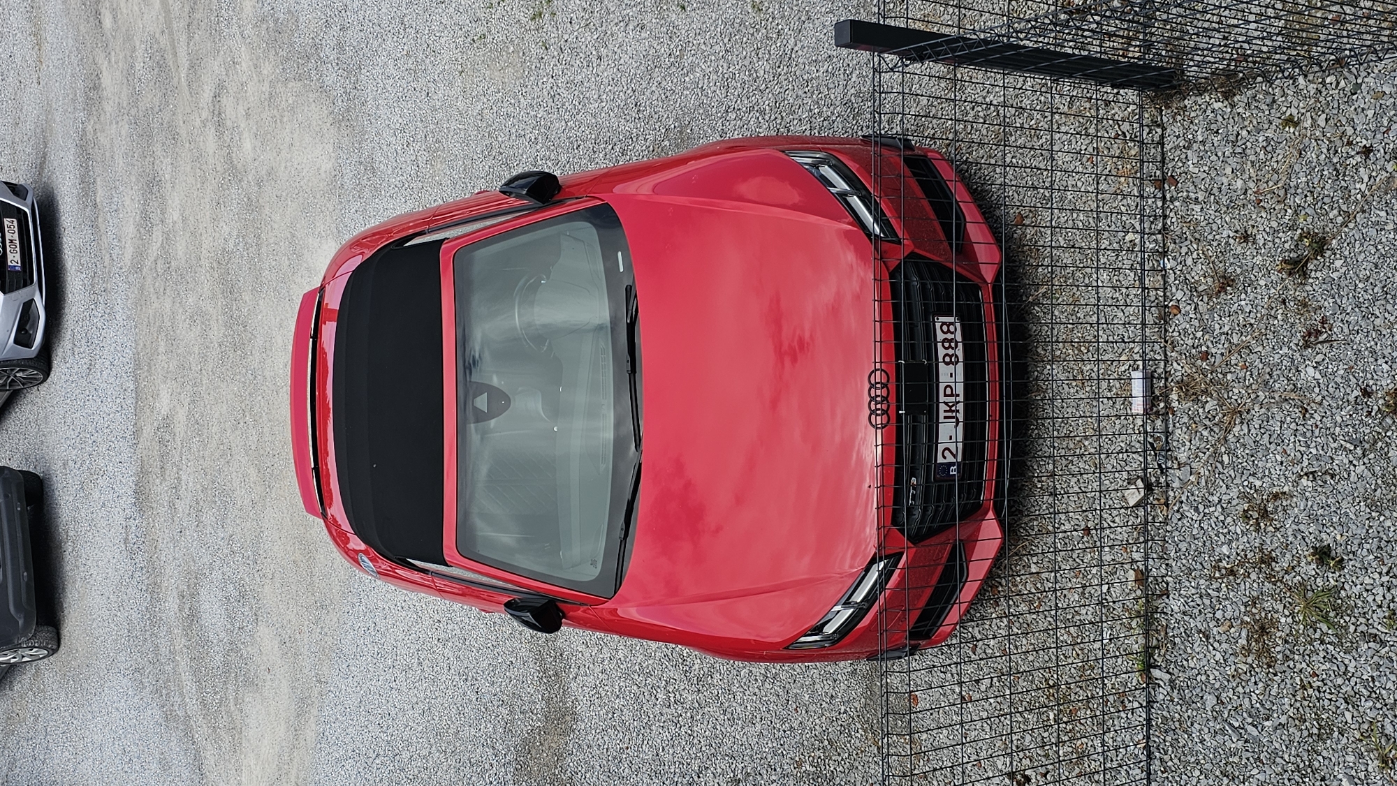 An overhead, slightly angled view captures a red Audi TT Roadster convertible parked on a light-colored gravel lot in Boussu-lez-Walcourt, Belgium. The vehicle's black soft top is in a retracted position, covered by a red boot. The front license plate, "2-JKP-888", bears a small 'B' indicating its Belgian registration. The Audi's front fascia features a large dark hexagonal grille with the Audi rings and modern LED headlights. Reflections of an overcast sky are visible on the car's paintwork and windshield. In the upper right background, a silver Audi, identifiable by its front grille and license plate "2-GOM-054", is partially visible. To the upper left, a portion of a dark-colored vehicle's rear bumper and wheel are also present. The foreground is occupied by a black metal mesh fence, through which sparse weeds and grass protrude from the gravel surface. No individuals are present in the scene.