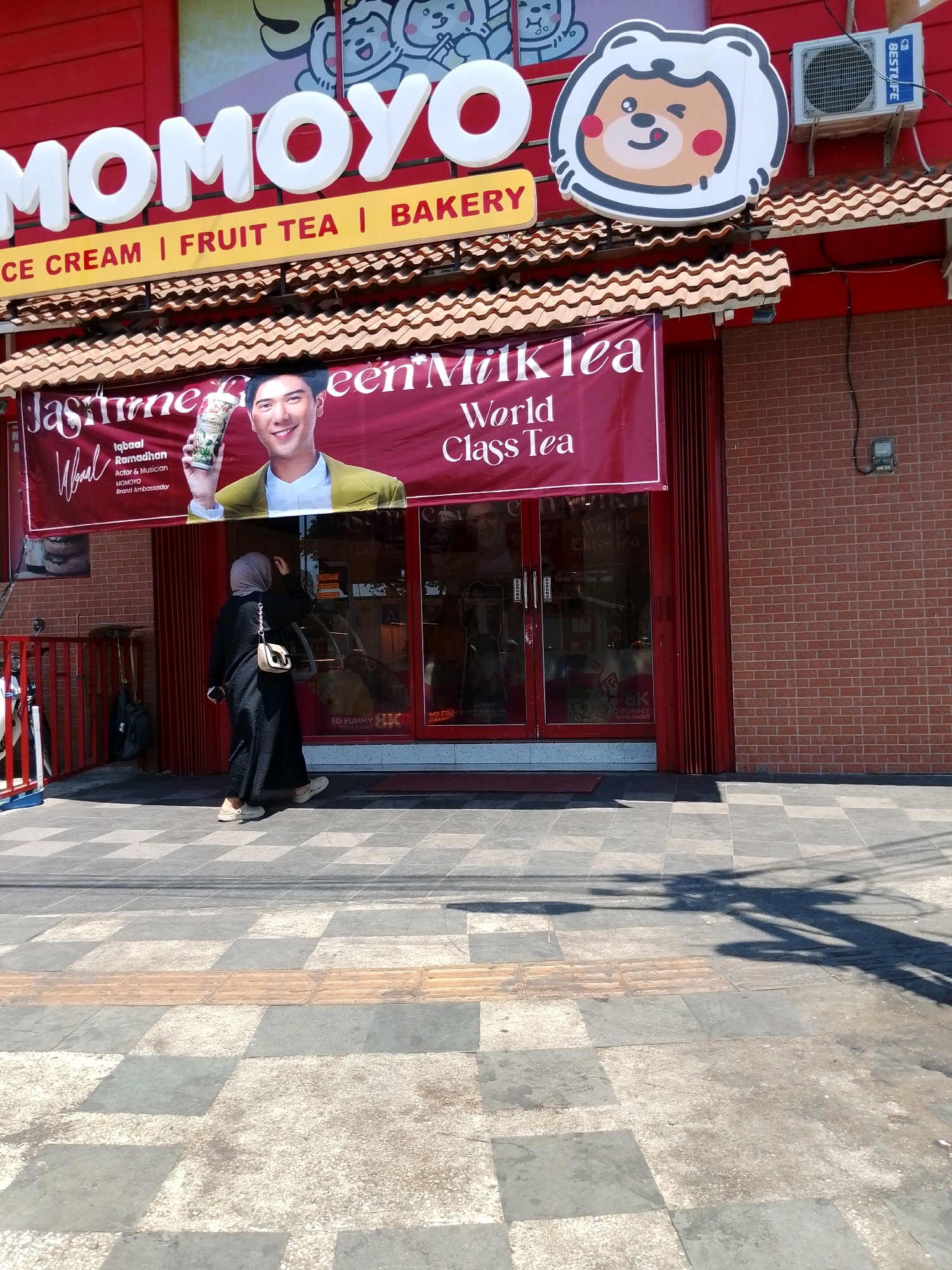 A storefront, identified by the prominent sign "MOMOYO," is visible, advertising "ICE CREAM | FRUIT TEA | BAKERY." A banner displays an advertisement for "Jasmine Green Milk Tea" featuring a man holding a drink, with the text "Iqbaal Ramadhan Actor & Musician MOMOYO Brand Ambassador" and "World Class Tea." The building's facade is painted red, with a tiled roof overhang. An air conditioning unit is mounted on the upper right wall. A woman wearing a black abaya and a grey hijab stands on the sidewalk in front of the store, facing away from the camera and adjusting her hijab. The sidewalk is paved with a checkerboard pattern of grey and white tiles, with tactile paving strips in a lighter brown color. A red metal fence runs along the left side of the sidewalk. The scene suggests a commercial area in the city of Pasuruan, Indonesia.