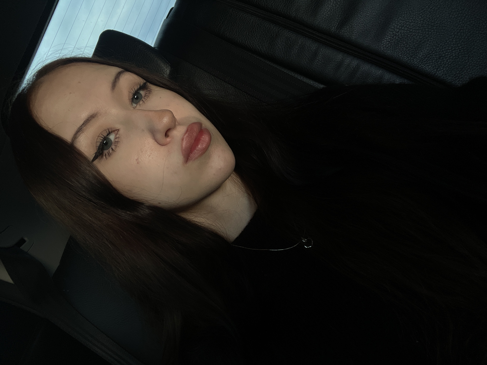 A young woman with long, dark brown hair and striking green eyes, adorned with sharp winged eyeliner, looks directly at the camera. Her lips are slightly parted, and she wears a delicate silver necklace with a small heart pendant. She is seated in the back of a dark leather car, with the headrest and seatbelt visible. The background suggests the car is moving, with a blurred view of the sky through the window. The overall lighting is dim, creating a moody and intimate atmosphere. The image appears to have been taken indoors, possibly during the evening or in a low-light environment.