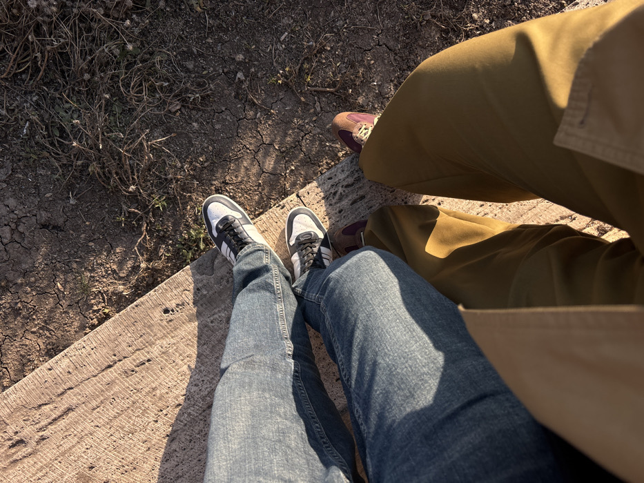 An overhead view depicts the lower extremities of two individuals positioned outdoors in Yerevan, Armenia. The subject on the left wears blue denim jeans and white and black athletic shoes with dark laces, while the subject on the right wears olive green trousers and brown and burgundy athletic shoes with light laces, with a light brown or beige coat visible at the bottom of the frame. Both subjects are positioned with their feet near the juncture of two distinct ground surfaces. To the upper left and central left, a textured, light-colored concrete or stone slab is visible. To the upper right and central right, the ground consists of dry, cracked earth with sparse, withered vegetation. Strong sunlight illuminates the scene, casting shadows across the surfaces and subjects' legs. No direct interaction between the subjects is discernible beyond their proximity.