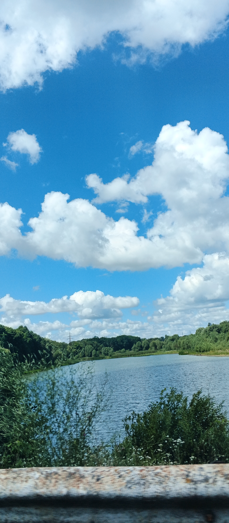 The image depicts a body of water, likely a lake or river, surrounded by lush green trees. The sky above is a vibrant blue, dotted with fluffy white clouds. In the foreground, a weathered, possibly metallic, railing or barrier runs horizontally across the bottom of the frame, slightly out of focus. The overall scene suggests a natural, tranquil environment. This particular view is situated in Kholodnianske, Ukraine, a location known for its natural landscapes and bodies of water.