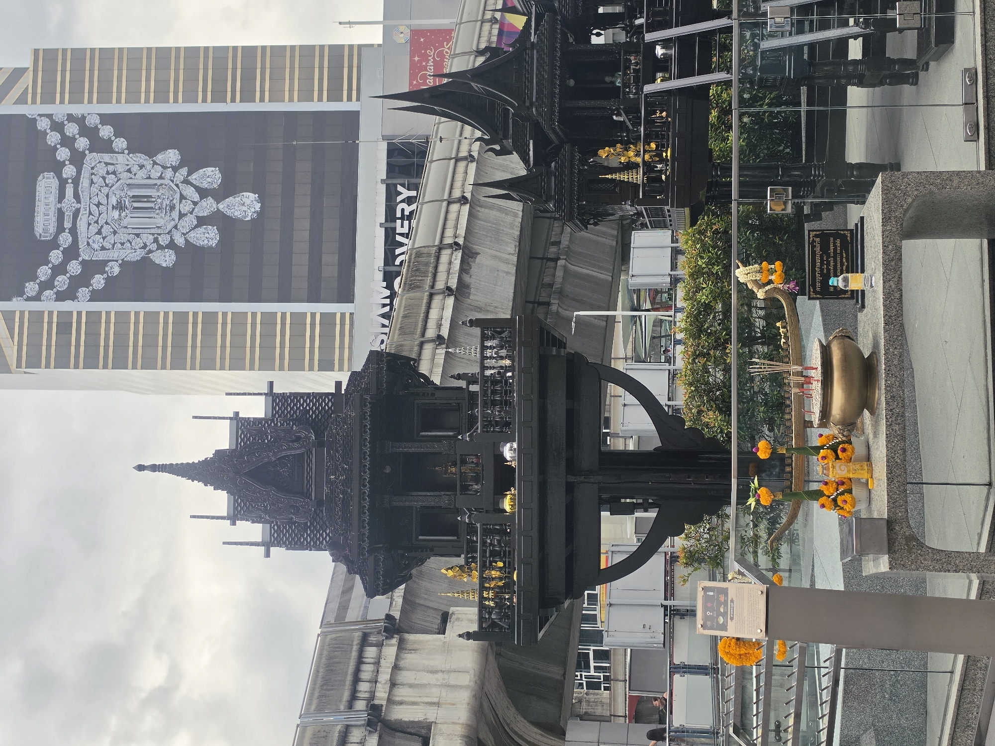 The image shows the Erawan Shrine, a famous shrine in Bangkok, Thailand, dedicated to the Hindu god Brahma. It is located at the Grand Hyatt Erawan Hotel intersection. The shrine is a small, ornate structure, decorated with flowers, incense, and other offerings. A sign containing the sacred religious mantra is visible. The photograph captures the reflection of the shrine in nearby windows, showing surrounding buildings, including the Siam Discovery shopping mall, as well as an image of diamonds on a building. The elevated BTS Skytrain tracks are also visible, indicating the shrine's proximity to the city's public transportation system.