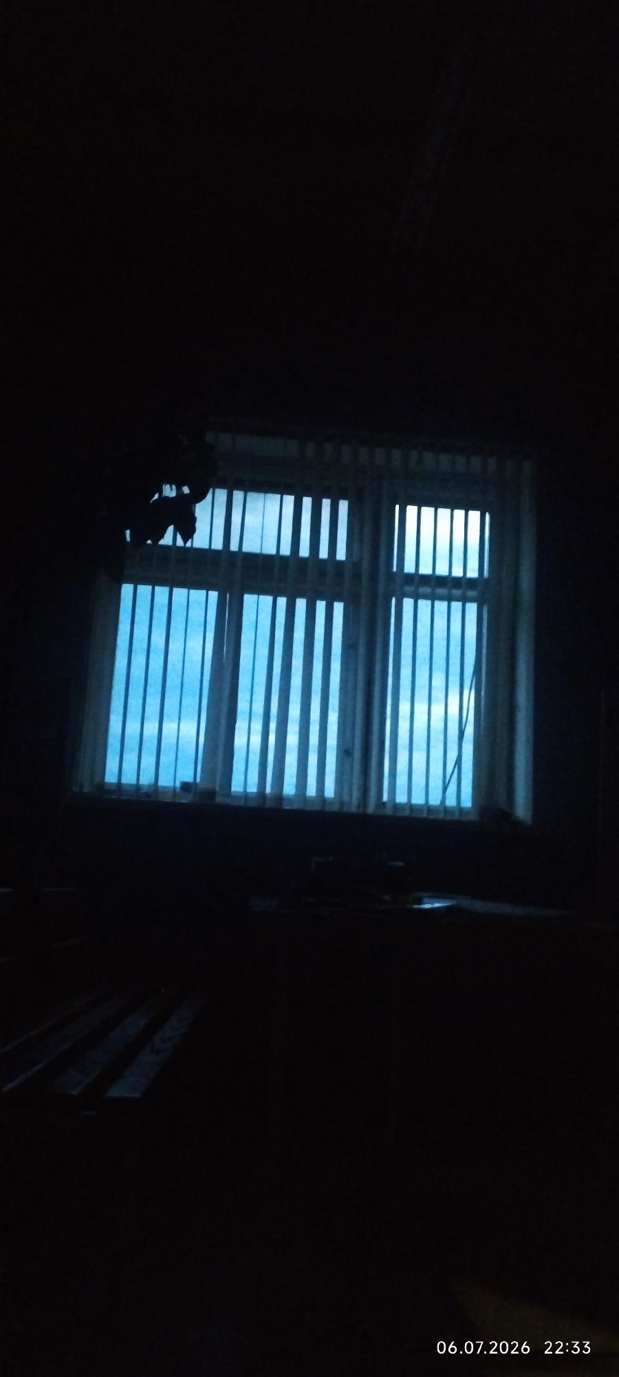 The image depicts an interior scene at night, with a window covered by vertical blinds that are partially open. The blinds reveal a bright blue sky with some clouds. The room itself is very dark, with only faint outlines of furniture visible. To the left of the window, a dark silhouette of what appears to be a hanging light fixture is visible. In the foreground, the edge of a wooden floor or furniture is dimly illuminated. The bottom right corner of the image shows a timestamp: "06.07.2026 22:33". The location is identified as Покосное, Russia.