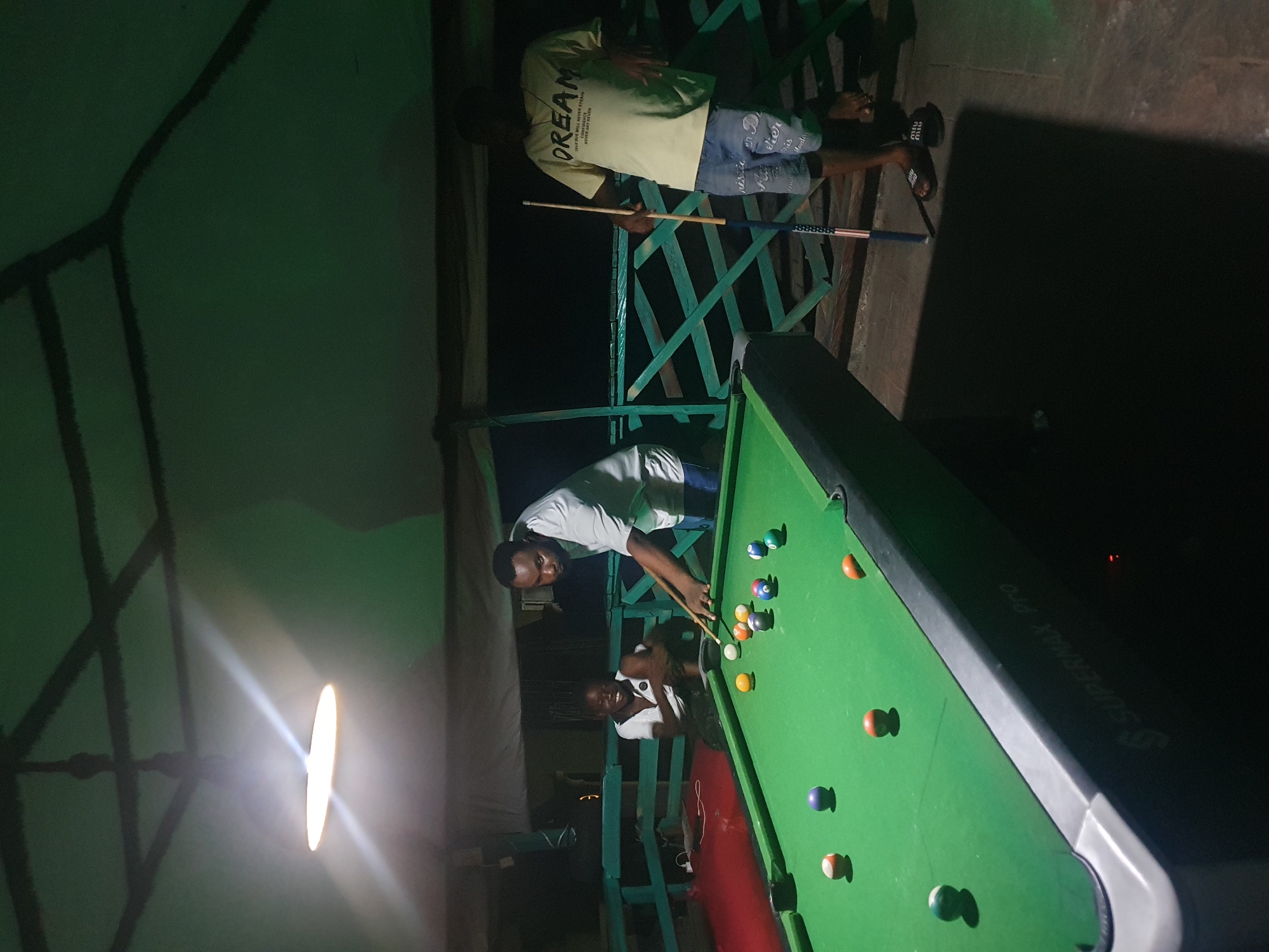 A group of people are playing billiards on a green pool table under a covered patio. A man in a white t-shirt is focused on aiming a shot with a cue stick. To his left, a young person in a white t-shirt watches with a wide smile. In the background, another man in a light yellow t-shirt with the word "DREAM" printed on it leans against a green wooden railing. The environment appears to be outdoors at night, possibly a backyard or an outdoor recreational area in Lokoja, Nigeria, indicated by the traditional Nigerian architectural elements of the covered patio and the lush green surroundings. The lighting is dim, provided by a single bulb overhead, casting long shadows. The pool table has several balls scattered across its surface, suggesting the game is in progress.