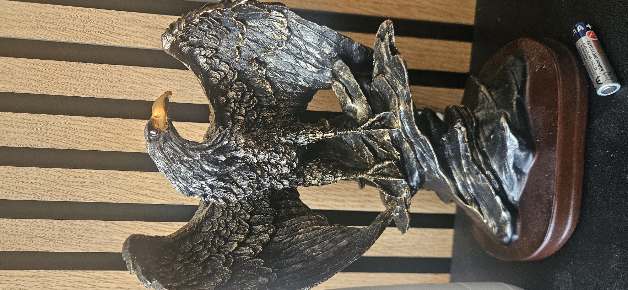 A figurine of an eagle with outstretched wings sits on a brown-and-black surface, next to a AAA battery, seemingly indoors. The background features light brown and black horizontal stripes. There is no information to determine the exact location within Spijkenisse, Netherlands.