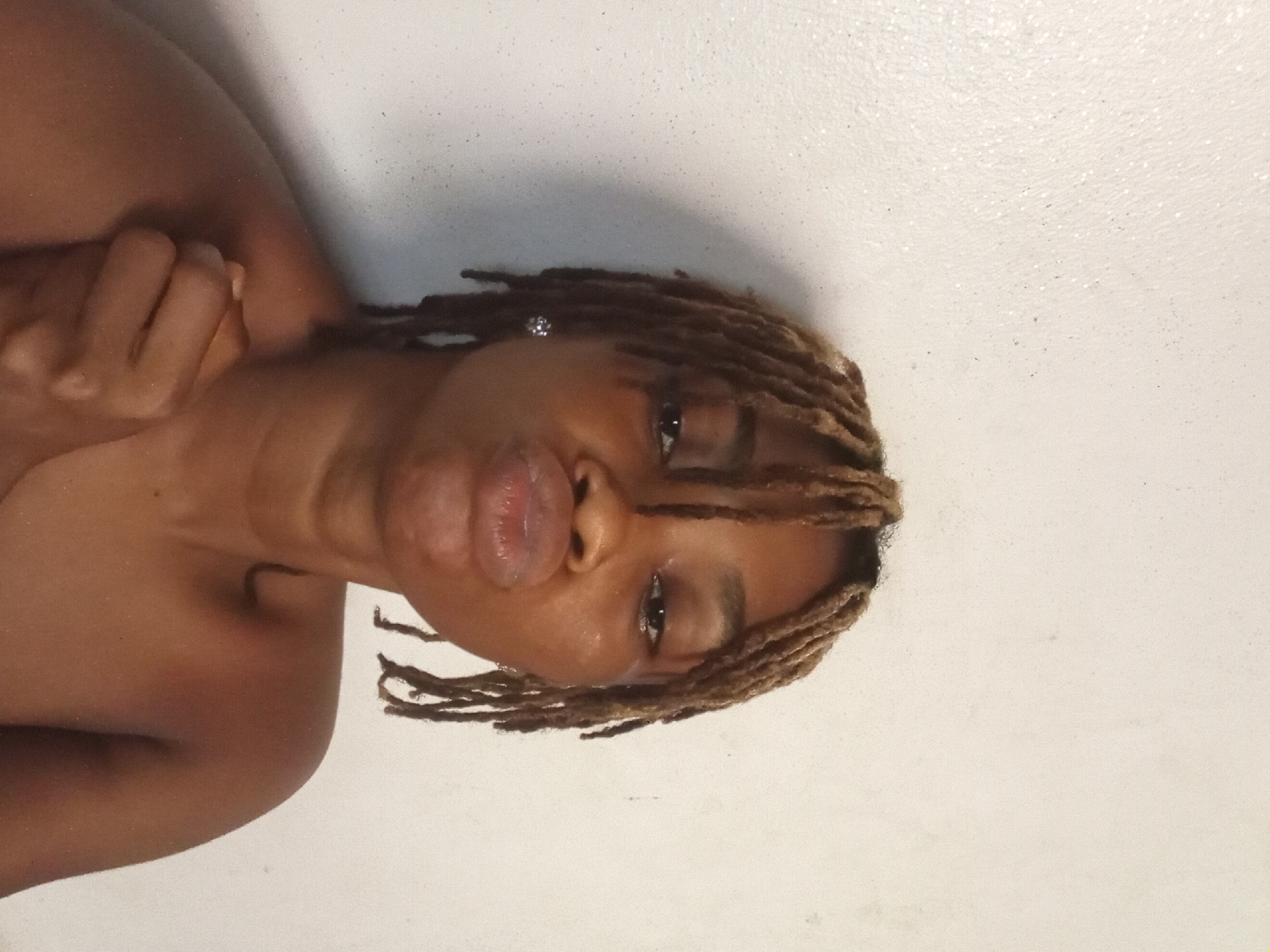 A woman with brown skin, short brown dreadlocks, and a silver earring in her hair is lying on a white surface, possibly a bathtub or bed. She is facing the camera, her eyes are open, and she is puckering her lips. Her left arm is bent and her hand is resting on her shoulder. The location is unknown, and there is no evidence to suggest that the image was taken in Shagamu, Nigeria.