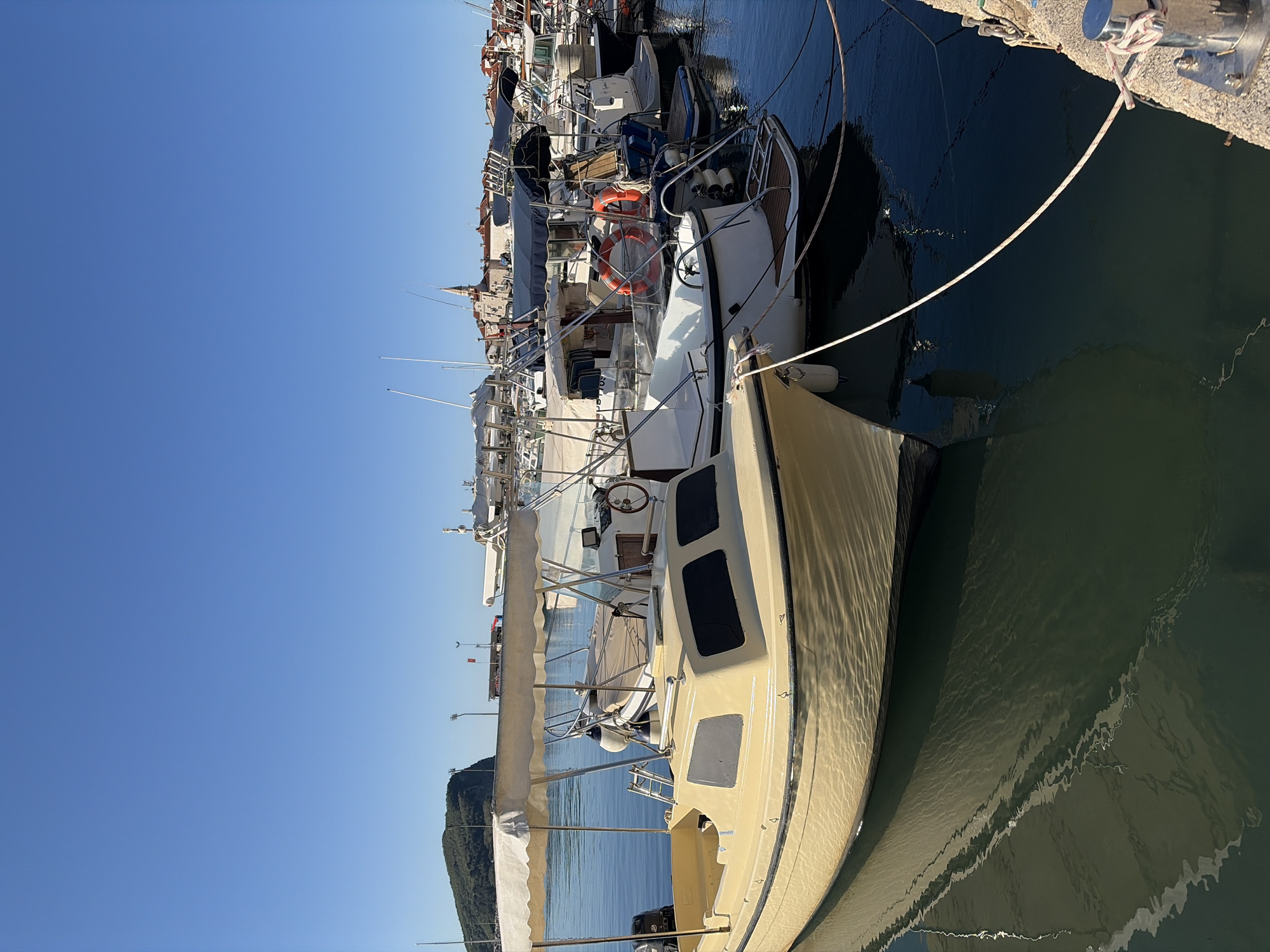 The image shows a marina scene in Budva, Montenegro on a clear, sunny day. Several boats are moored along a concrete pier. A cream-colored sailboat with a mast and rigging is prominent in the foreground, tied to the pier with white ropes. The water reflects rippling patterns of light. In the background, other boats of varying sizes are visible, creating a dense array. The skyline of Budva, with buildings and a church steeple, is visible beyond the boats. A green, hilly landscape is also visible in the distance.
