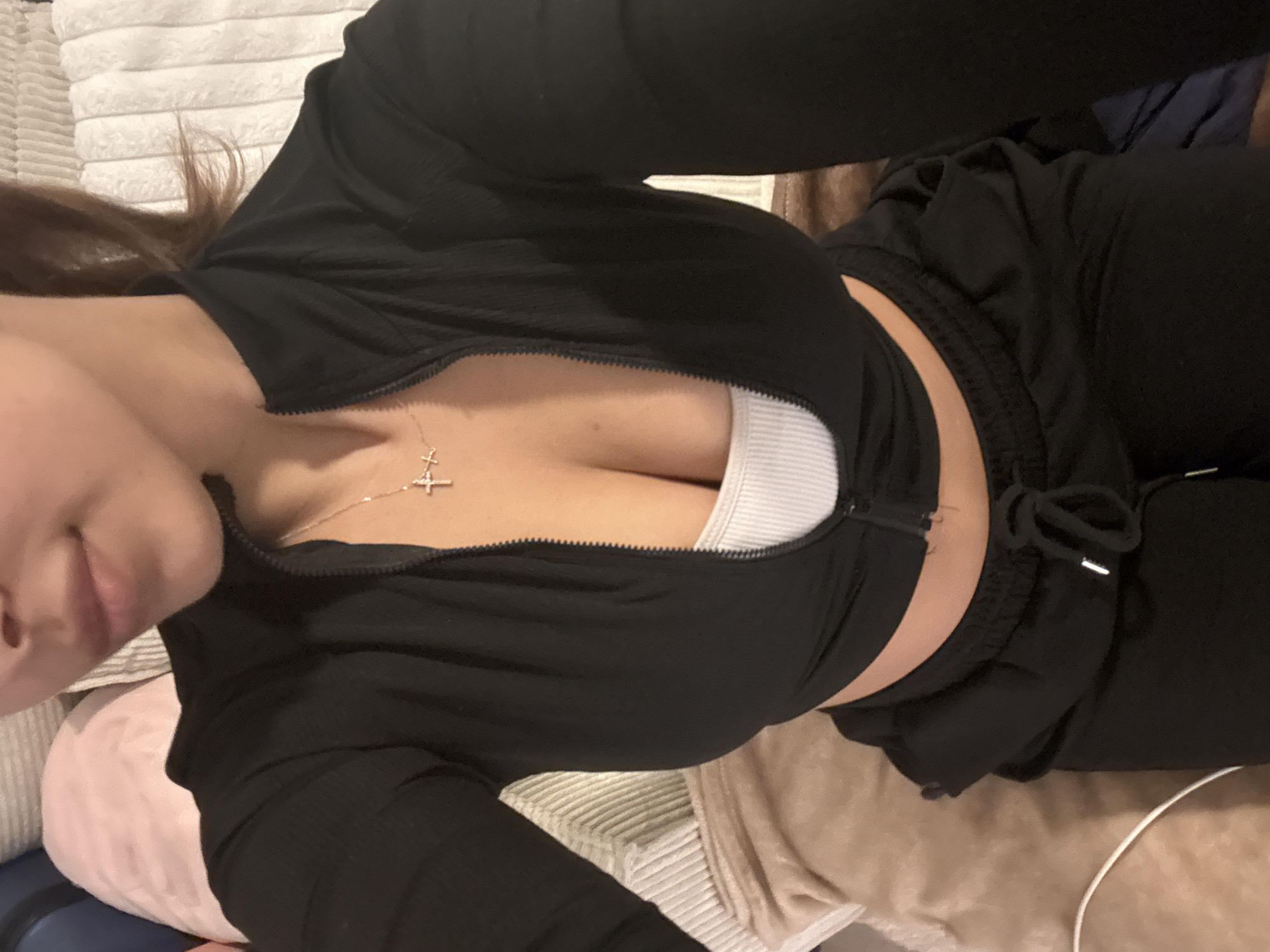 A woman is lying down, possibly on a bed, wearing a black zip-up jacket over a white tank top, and black sweatpants. She is also wearing a necklace with a cross pendant. The background includes various pillows and blankets. There is no indication of Mönchengladbach, Germany.