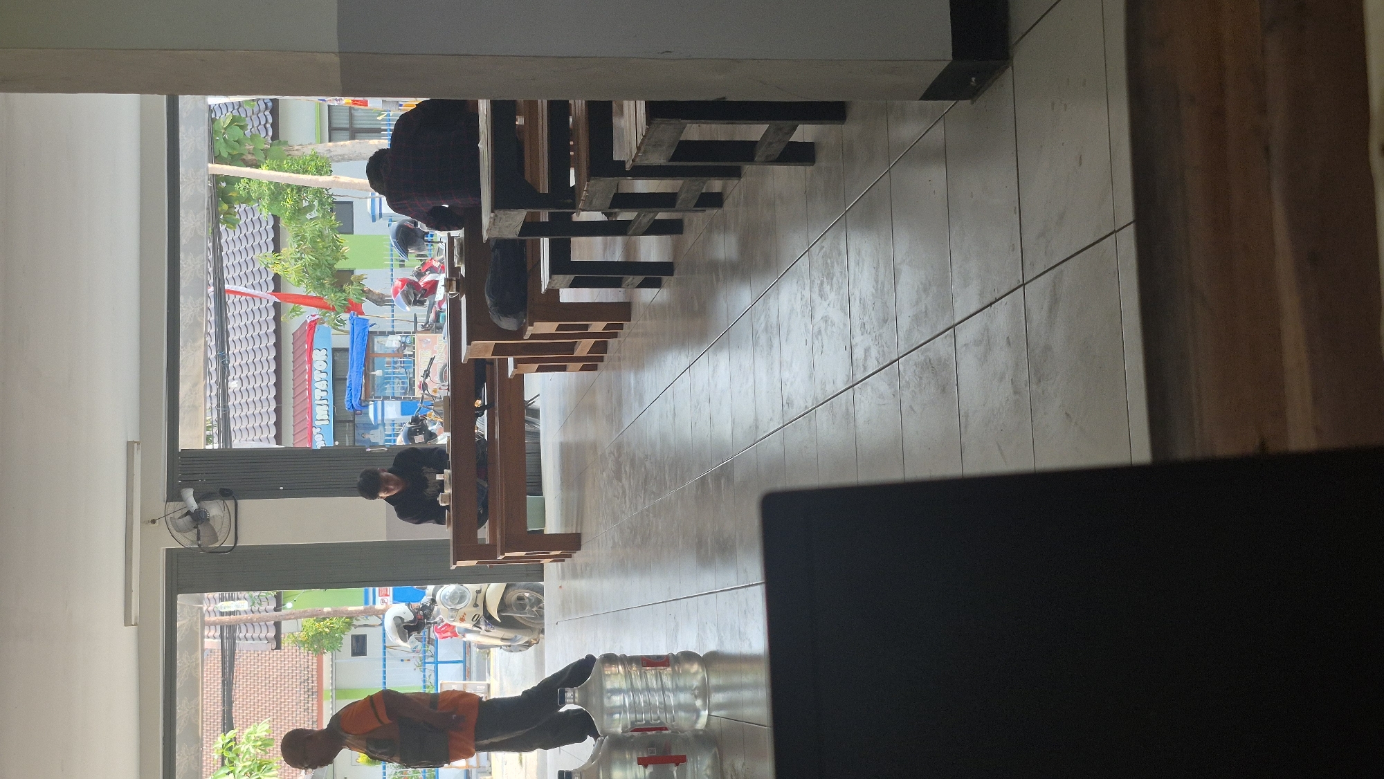 The image depicts an indoor scene with an outdoor view through large windows. The interior features tiled flooring and wooden furniture, including tables and benches. Two large water jugs are visible near the left side of the frame. In the foreground, the edge of a dark object, possibly a laptop, is present.
Several individuals are present. On the left, a person wearing an orange vest and dark pants stands facing left. In the middle, a person in a black hoodie is seated at a wooden table, looking down. Further to the right, another person, seen from the back, is wearing a red and black checkered shirt and is seated at a wooden table.
Outside the windows, a street scene is visible. There are buildings with tiled roofs, trees, and several motorcycles parked. A sign that reads "IMITATOR" is visible on one of the buildings. A red and white flag is also displayed.
The scene appears to be a public space, possibly a cafe or a communal area. The overall lighting suggests daytime. The location is identified as Bojonegoro, Indonesia, based on the provided context.
