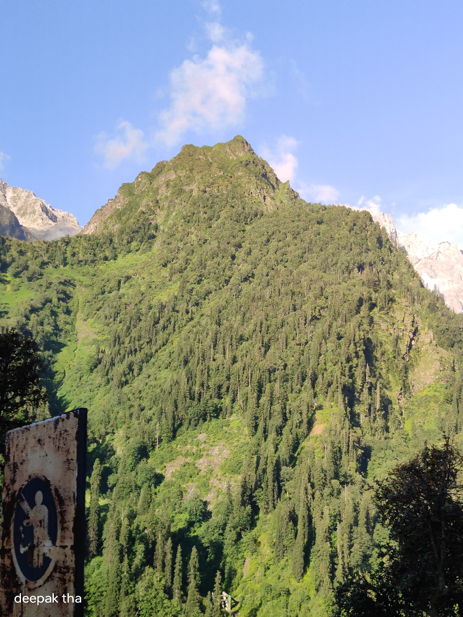 This is a full-frame, eye-level, outdoor, vertical shot of a lush green mountain in Burwa, India. The mountain is densely covered with a variety of trees, with a prominent peak at the top. The sky above is a clear blue with scattered white clouds. In the lower left corner of the image, there is a rusty, partially obscured sign with a circular emblem depicting a person holding a rifle. Below the sign, the text "deepak tha" is visible. The lighting suggests it is daytime, with the sun illuminating the mountain and sky.