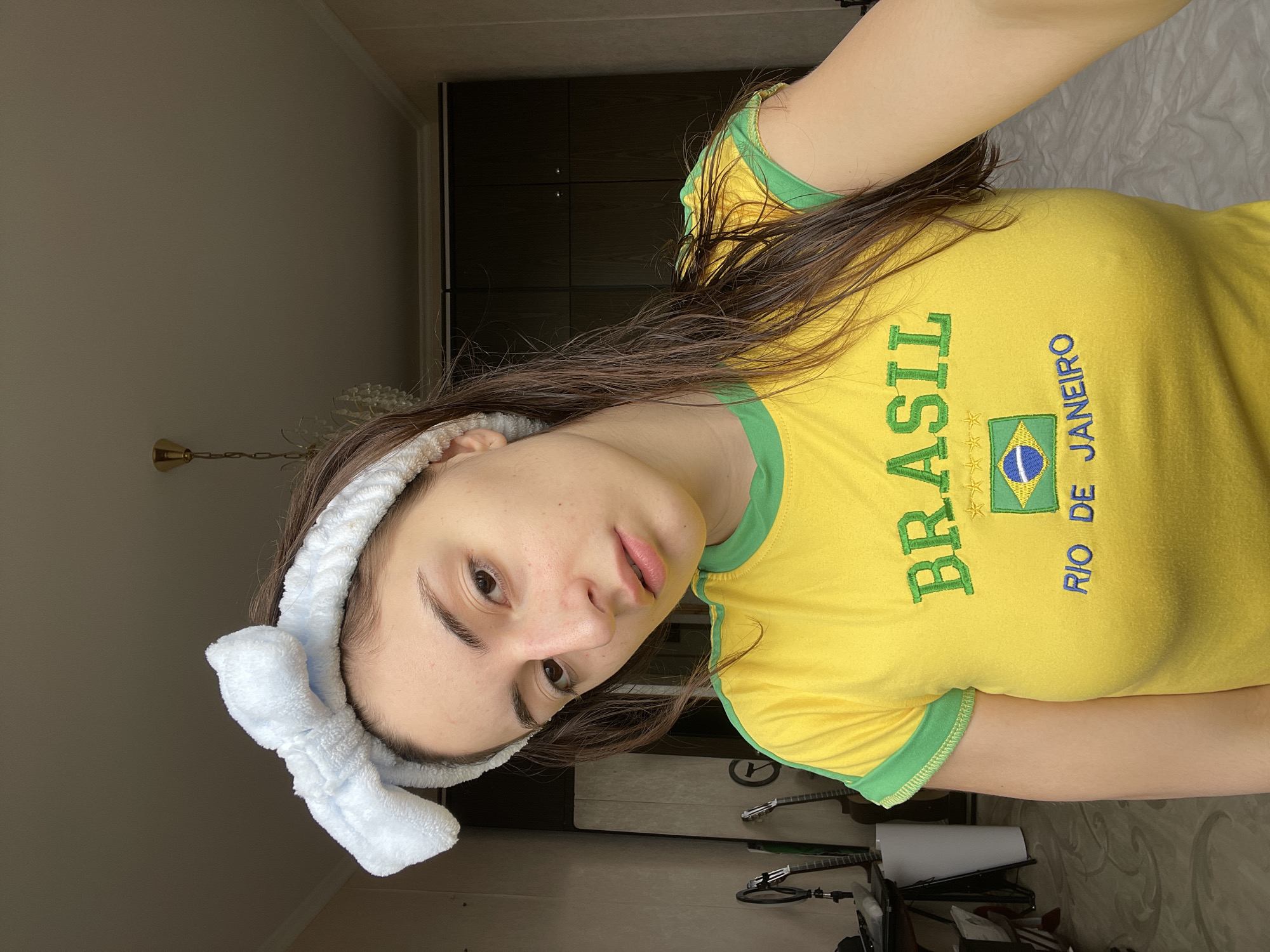 A young person with long, dark brown hair, appearing to be female, is depicted in an indoor setting in Moscow, Russia. The individual is wearing a light blue, fluffy headband with a bow, securing their hair back, and a vibrant yellow t-shirt with green trim. The t-shirt features green embroidered text "BRASIL" vertically, and blue embroidered text "RIO DE JANEIRO" horizontally, alongside a small embroidered Brazilian flag with its motto "ORDEM E PROGRESSO" visible.
The person is looking directly at the camera with a neutral expression. The background reveals a simple room with light-colored walls. To the right, dark wooden cabinets or a wardrobe are visible. Below, glimpses of musical instruments, possibly guitars on stands, and a patterned rug or carpet can be seen on the floor. A decorative light fixture or chandelier is partially visible on the left wall. The lighting suggests natural daylight, possibly diffused or from an overcast sky. The image is taken in a casual, home environment, possibly a bedroom or living area. The overall scene is calm and unposed, hinting at a moment of leisure or personal care given the headband.