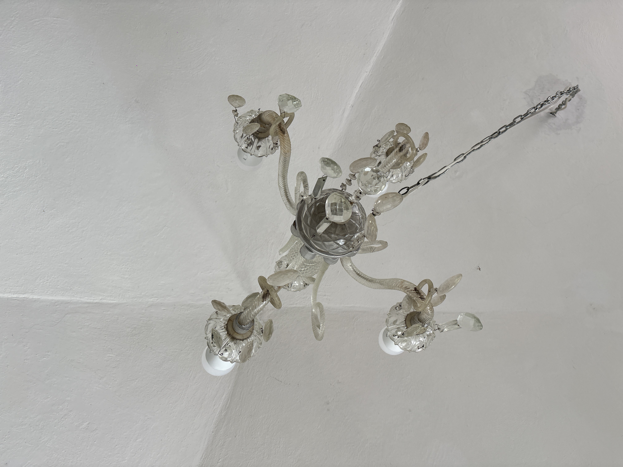 A low-angle shot depicts a white, plastered ceiling with a crystal chandelier suspended from a chain. The chandelier has a central, faceted crystal body with four arms branching out, each culminating in a bulb surrounded by decorative crystal elements. The ceiling shows subtle texture and a slight vaulting, suggesting an older construction style. The location is Puebla, Mexico.