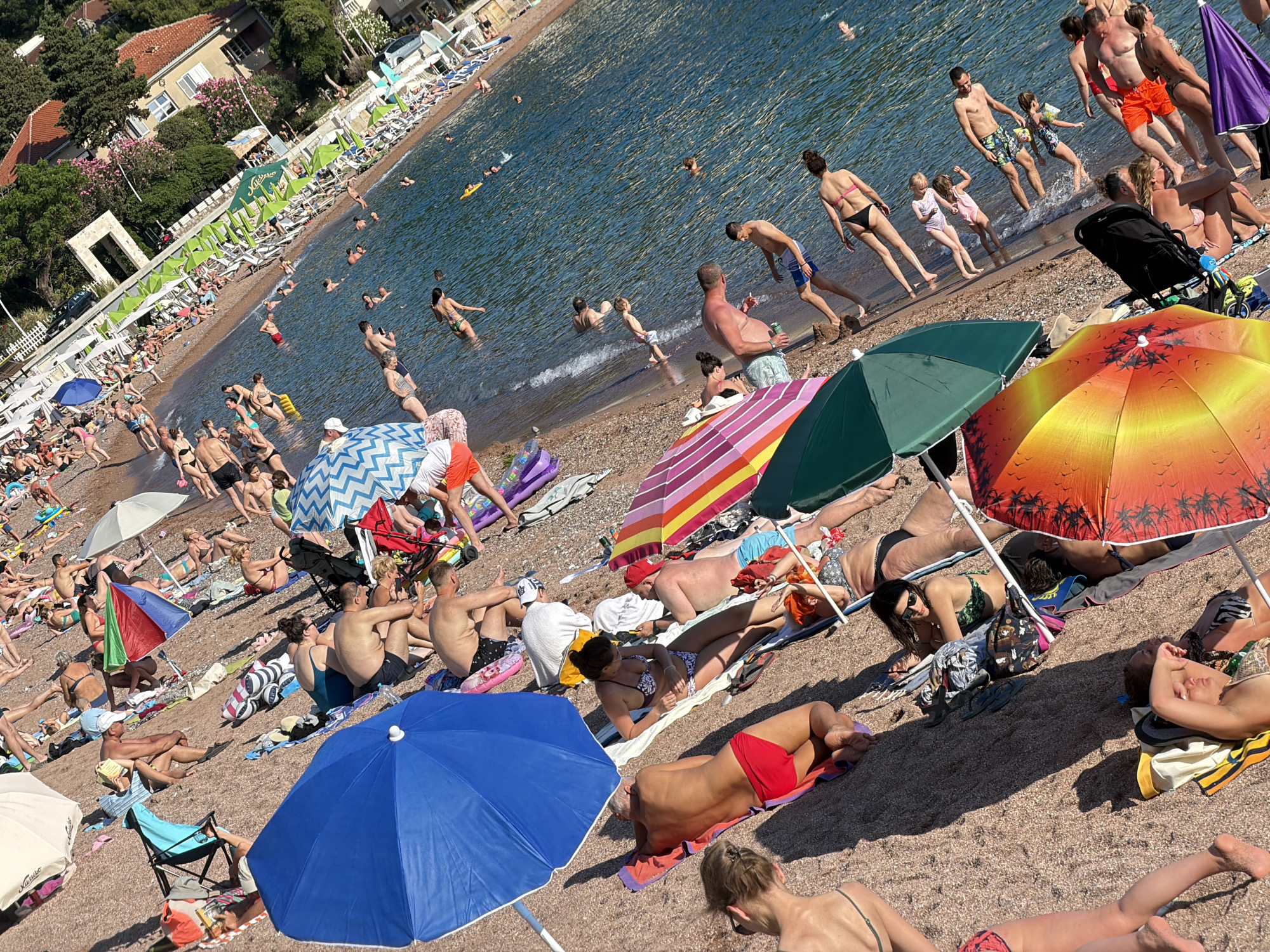 The image depicts a crowded beach scene in Petrovac centar, Montenegro. Numerous people are sunbathing on the sand, lying on towels or beach chairs. Some individuals are in the water, swimming or wading. Several beach umbrellas provide shade. In the background, buildings and lush greenery are visible, indicating a coastal town setting. The overall atmosphere is one of leisure and recreation.