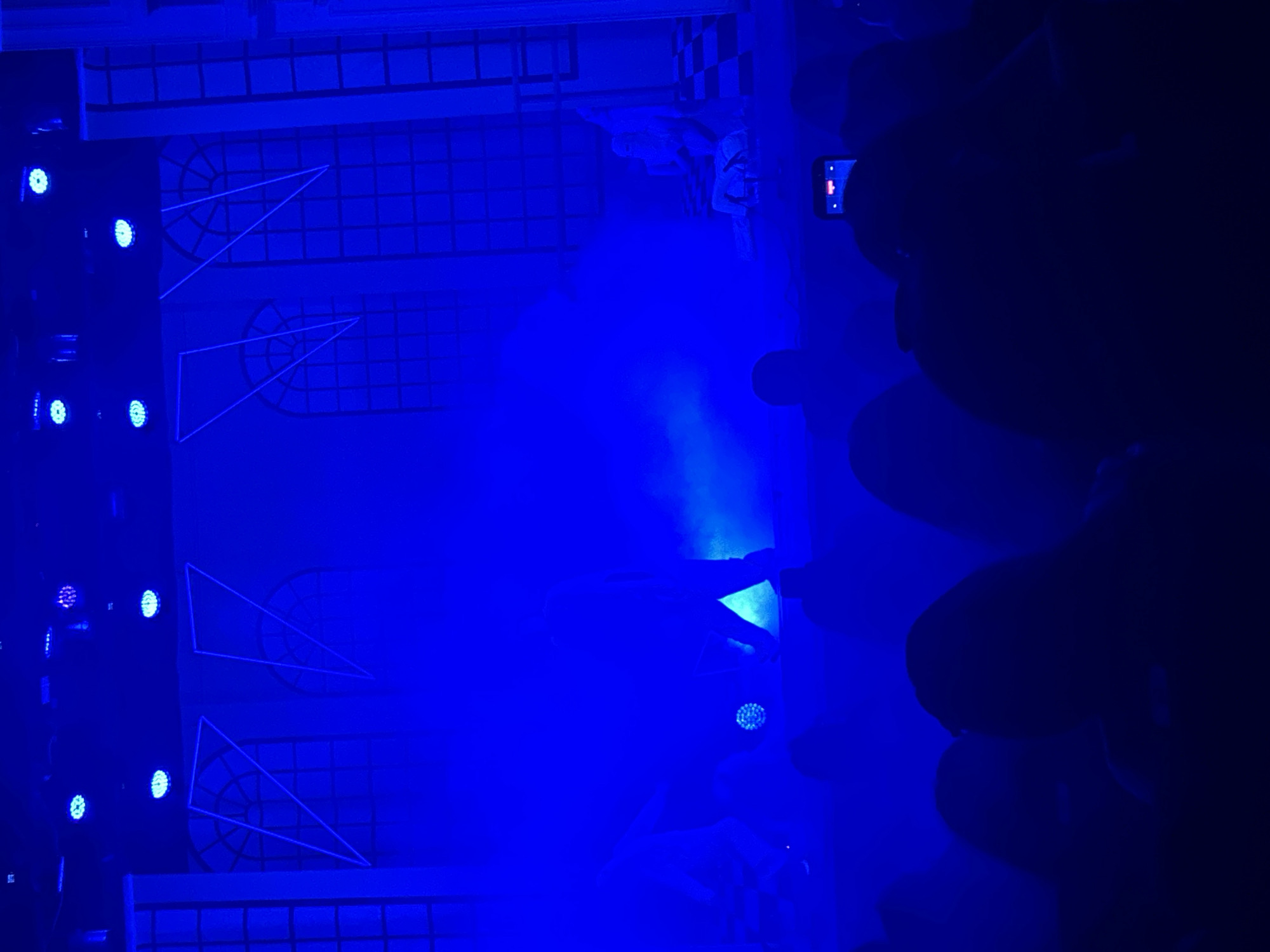 The image captures a scene bathed in a vibrant blue light. The background consists of architectural elements featuring geometric shapes, grid patterns, and circular designs, suggesting a theatrical or performance setting. On the right side of the frame, a crowd of people is visible, their forms largely silhouetted, with one person holding up a smartphone. The image's orientation suggests the scene is viewed from the side, capturing a dynamic performance moment. Without any additional information, I can not determine the specific location in Sofia, Bulgaria.