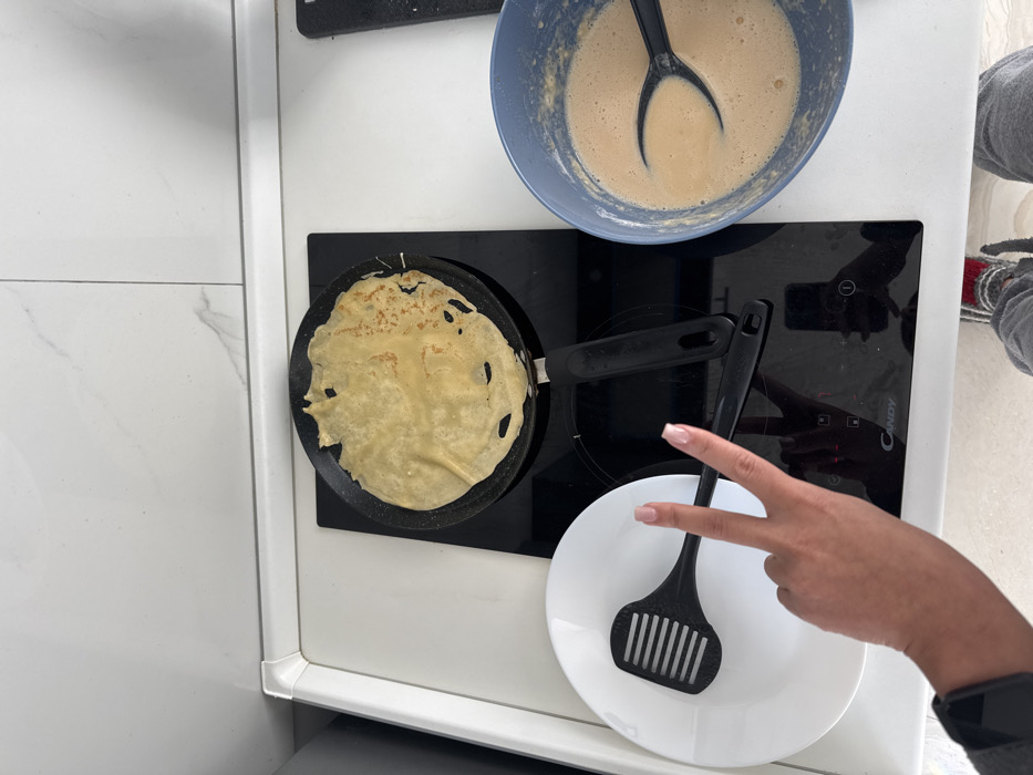 This image captures an overhead view of a kitchen countertop in Budva, Montenegro, depicting the preparation of pancakes or crepes. A white countertop with a white tiled backsplash serves as the backdrop. Centrally located on the countertop is a black "Candy" brand induction or ceramic stovetop. A black frying pan rests on the stovetop, containing a partially cooked, light yellow pancake with some browned, irregular edges. To the left of the stovetop, a white round plate holds a black slotted spatula. A human hand, featuring light-colored manicured fingernails, is positioned in the lower-left foreground, forming a "V" sign with the index and middle fingers. To the right of the stovetop, a blue mixing bowl holds a quantity of light brown liquid batter, with a black utensil immersed within it. Faint reflections of a person's legs/feet and what appears to be a smartphone are visible on the stovetop's shiny black surface.