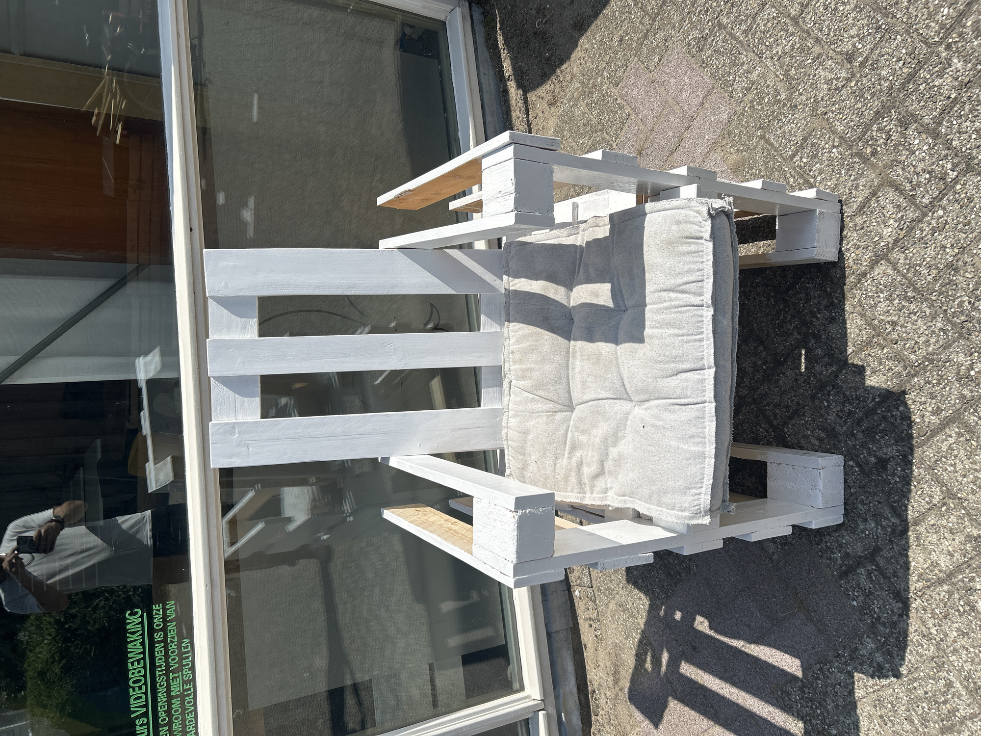 In Lisse, Netherlands, an outdoor scene features a white wooden chair with a gray cushion. The chair appears to be constructed from pallets and is positioned on a paved surface with visible brick patterns. Behind the chair is a large window, reflecting sunlight and showing a partial view of the interior. The reflection in the window includes a person holding a phone and text in Dutch, which translates to "ours VIDEO SURVEILLANCE. During opening hours, our store is not responsible for valuable items." The environment suggests it is a sunny day, with shadows cast on the ground.