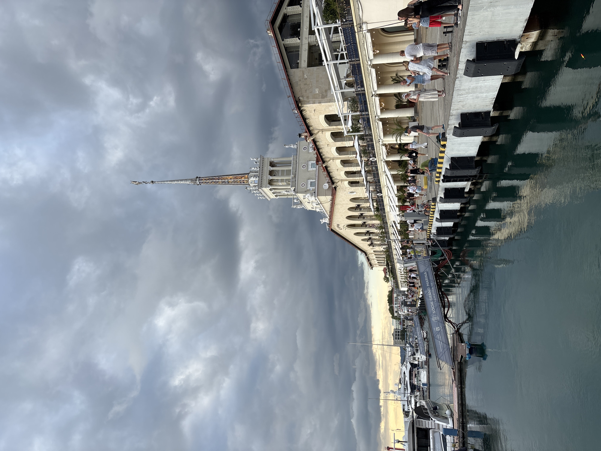 The image depicts a waterfront scene in Sochi, Russia, likely at the Sochi Grand Marina, as indicated by text on a gangway. The architecture of the building along the waterfront is distinctive, featuring arched colonnades and a tall, ornate tower with a spire. The sky is overcast with dramatic, dark clouds.
In the foreground, the dark water of the marina reflects the building and the sky. Several yachts and boats are docked in the harbor, some appearing to be large and luxurious. A gangway with "SOCHI GRAND MARINA" and "sgmarina.ru" visible connects to the dock.
Along the dock, a number of people are present. Some are walking, while others are standing and observing the surroundings. One person is fishing from the dock. The people are casually dressed, suggesting a daytime activity. The overall atmosphere appears to be a blend of bustling activity and the impending mood of an overcast sky.