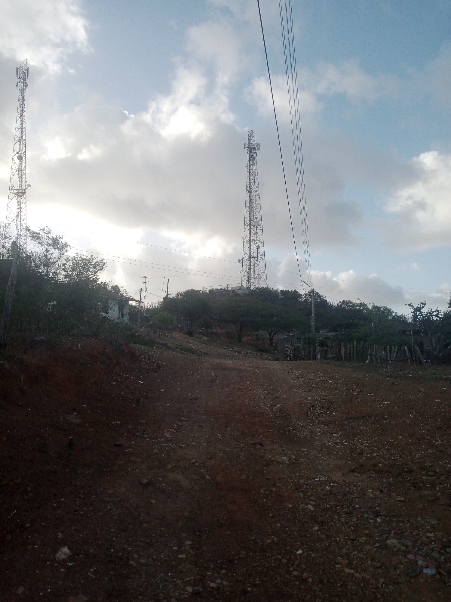The image captures an outdoor scene on a slightly overcast day. In the background, two tall, metallic cell towers stand prominently against a sky dotted with clouds. One tower is positioned on the left side of the frame, and another is further to the right, appearing slightly shorter in the distance. Several power lines traverse the upper portion of the image, stretching diagonally from the right.
The middle ground features a hillside covered with dense, green vegetation. A small, light-colored building with a green roof and barred windows is visible amidst the foliage. A rustic wooden fence runs along the slope. Various trees and shrubs are scattered across the hillside.
The foreground consists of a dirt road or path, characterized by its reddish-brown soil and scattered stones and pebbles. The ground appears somewhat uneven. The overall impression is of a rural or semi-rural setting, possibly on the outskirts of a town. The location is identified as La Tagua, Venezuela.