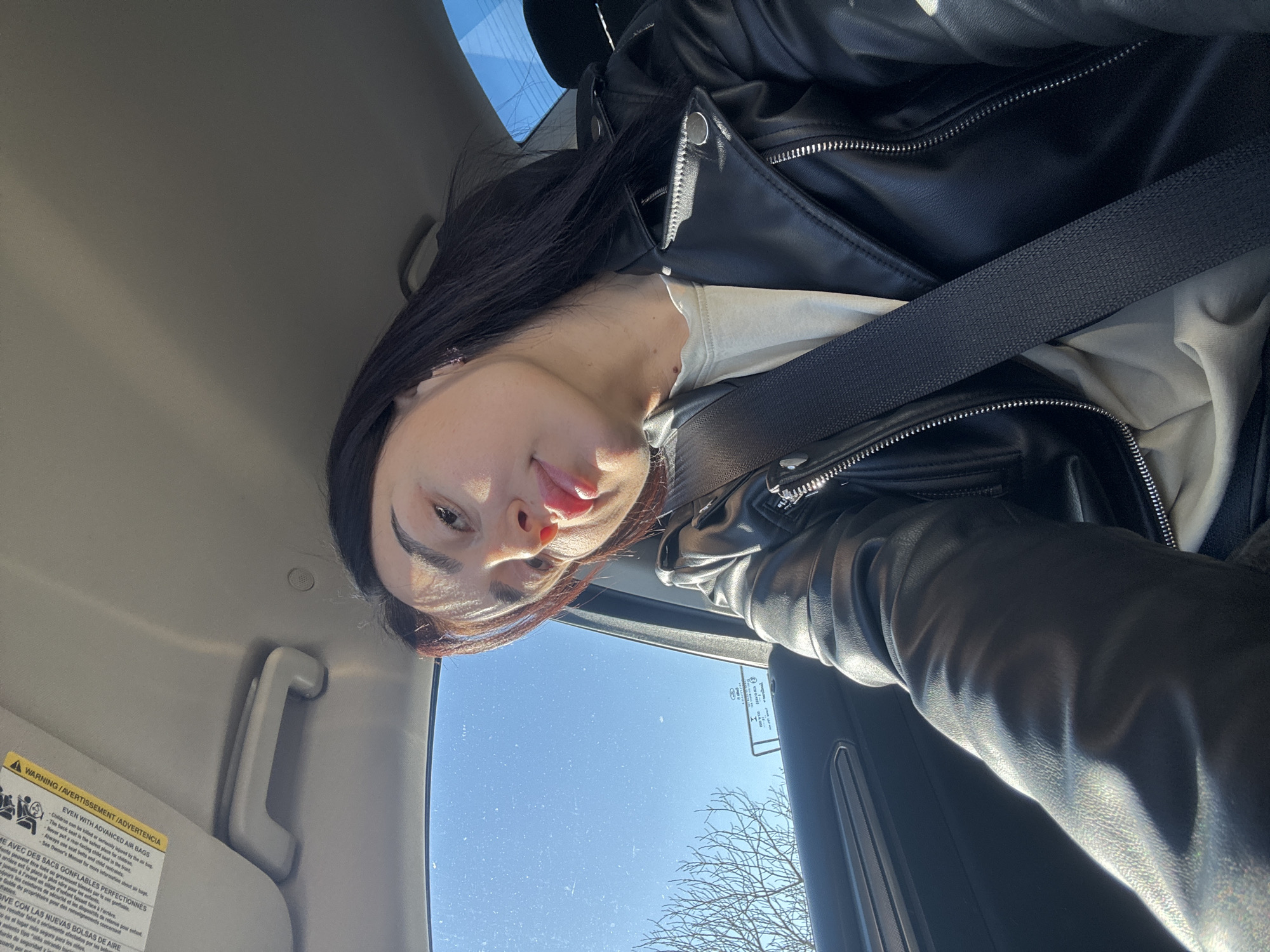 A young woman with long, dark hair, a black leather jacket, and a white shirt is in a car, wearing a seatbelt. The car appears to be in motion on a sunny day with a clear blue sky, visible through the window. An obscured tree branch is also visible through the window. An instruction label regarding airbags is visible in the upper left. There is no information on the exact location within Tbilisi, Georgia.