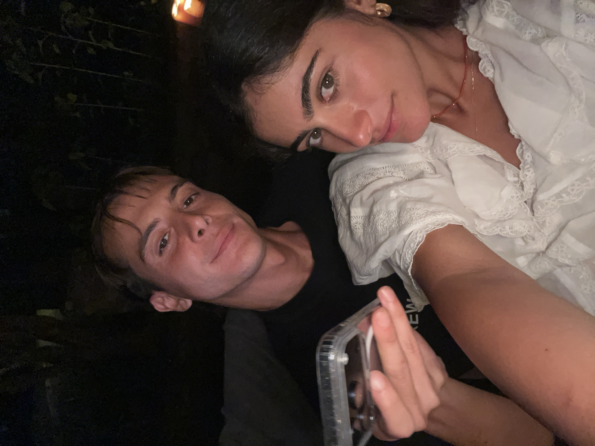 This is a selfie taken outdoors at night, possibly in the city of Vila Nova de Cerveira, Portugal. A young man in a black t-shirt is in the foreground on the left, holding up a clear phone case with a phone inside. He is smiling at the camera. To his right is a young woman wearing a white lace top. She has dark hair, prominent eyebrows, and is wearing gold hoop earrings and a delicate necklace. She is looking at the camera with a slight smile. In the background, out of focus, there are some tall, thin plants and two lit candles on a surface, suggesting an outdoor setting with ambient lighting. The overall scene appears to be casual and intimate.