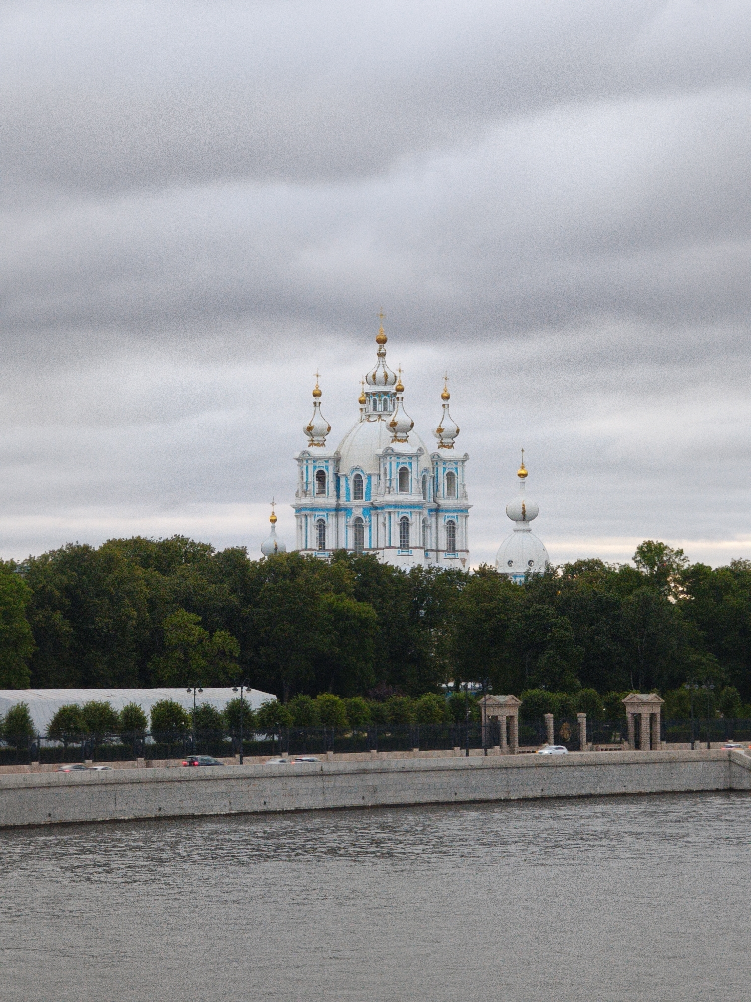 A wide shot shows the Smolny Cathedral in St. Petersburg, Russia, under a cloudy sky. The cathedral, a large white and blue building with gold domes, is surrounded by lush green trees. In the foreground, a river flows, with a concrete embankment and a road with cars on it. The scene is serene and picturesque, capturing the grandeur of the cathedral and its natural surroundings.
