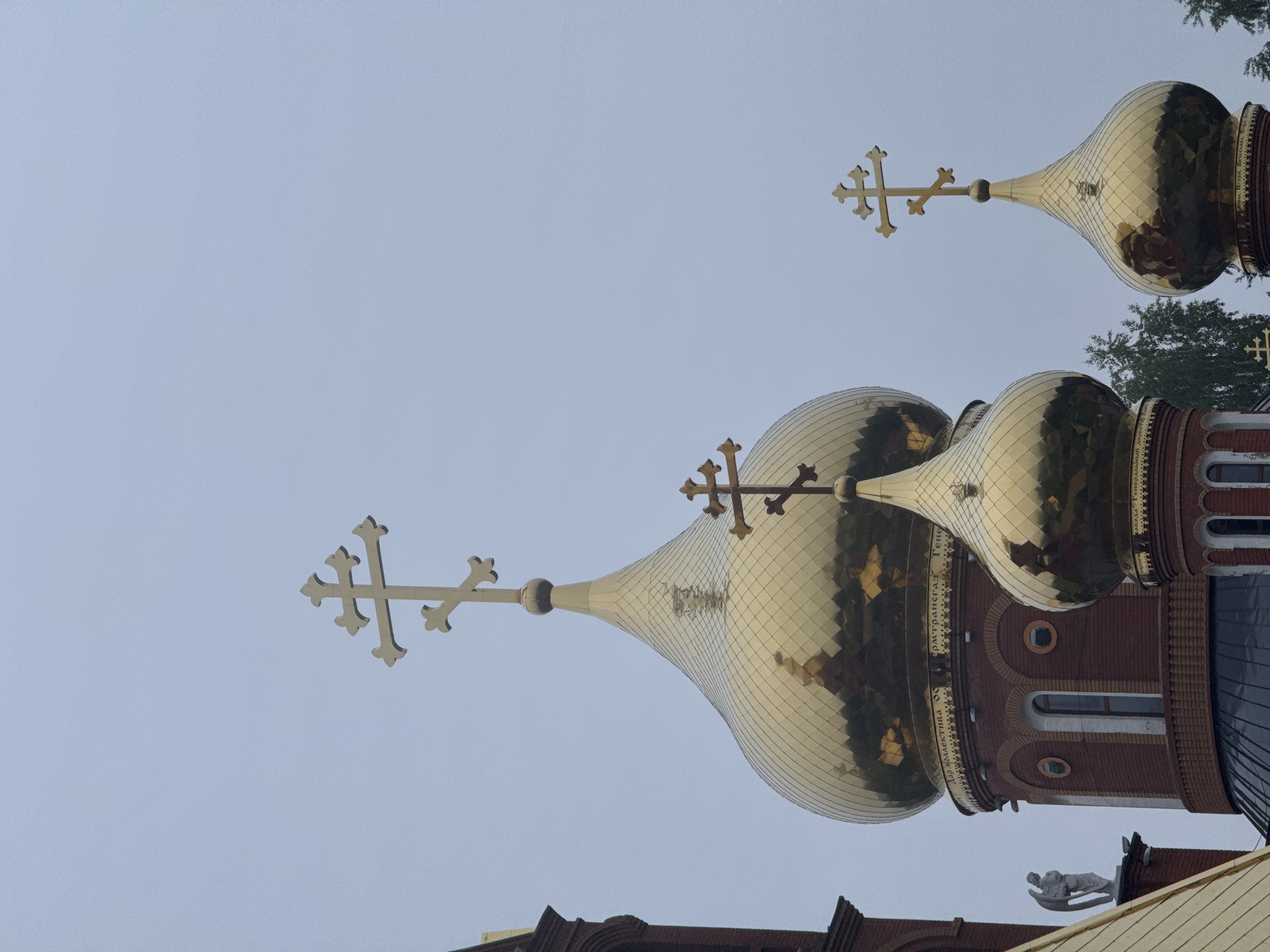 An image, presented rotated 90 degrees clockwise from its natural orientation, depicts the upper section of an Orthodox church against an overcast sky. When viewed upright, the scene prominently features multiple golden onion domes and intricate Orthodox crosses.
The central and largest dome is a vibrant gold, composed of many reflective tiles, giving it a textured, shimmering appearance. It tapers upwards to a slender golden spire, topped with a multi-barred Orthodox cross featuring distinctive trefoil ends. Around the cylindrical drum supporting this main dome, a clear Cyrillic inscription in black letters reads "БЛАГОСЛОВЕНЪ ЕСИ ГОСПОДИ" ("Blessed art Thou, O Lord"), followed by further, less legible text.
To the right of the central dome, a second, slightly smaller golden onion dome is partially visible, also adorned with a similar Orthodox cross. Further into the background, towards the upper right corner, another small golden dome and cross are faintly discernible.
The church structure itself, visible beneath the domes, is constructed from red brick, showcasing arched window frames and decorative elements. On a lower roofline section to the bottom right, a small white statue, possibly an angel or a religious figure, is perched. Hints of green tree foliage are visible on the right side and in the gaps between the architectural elements.
The sky is a uniform, soft light blue-grey, indicating an overcast or hazy daytime. The diffused lighting suggests no direct sun, possibly late morning or early afternoon. The architectural style, including the onion domes and Orthodox crosses, is characteristic of Russian Orthodox churches, aligning with the location context of Chaikovsky, Russia.