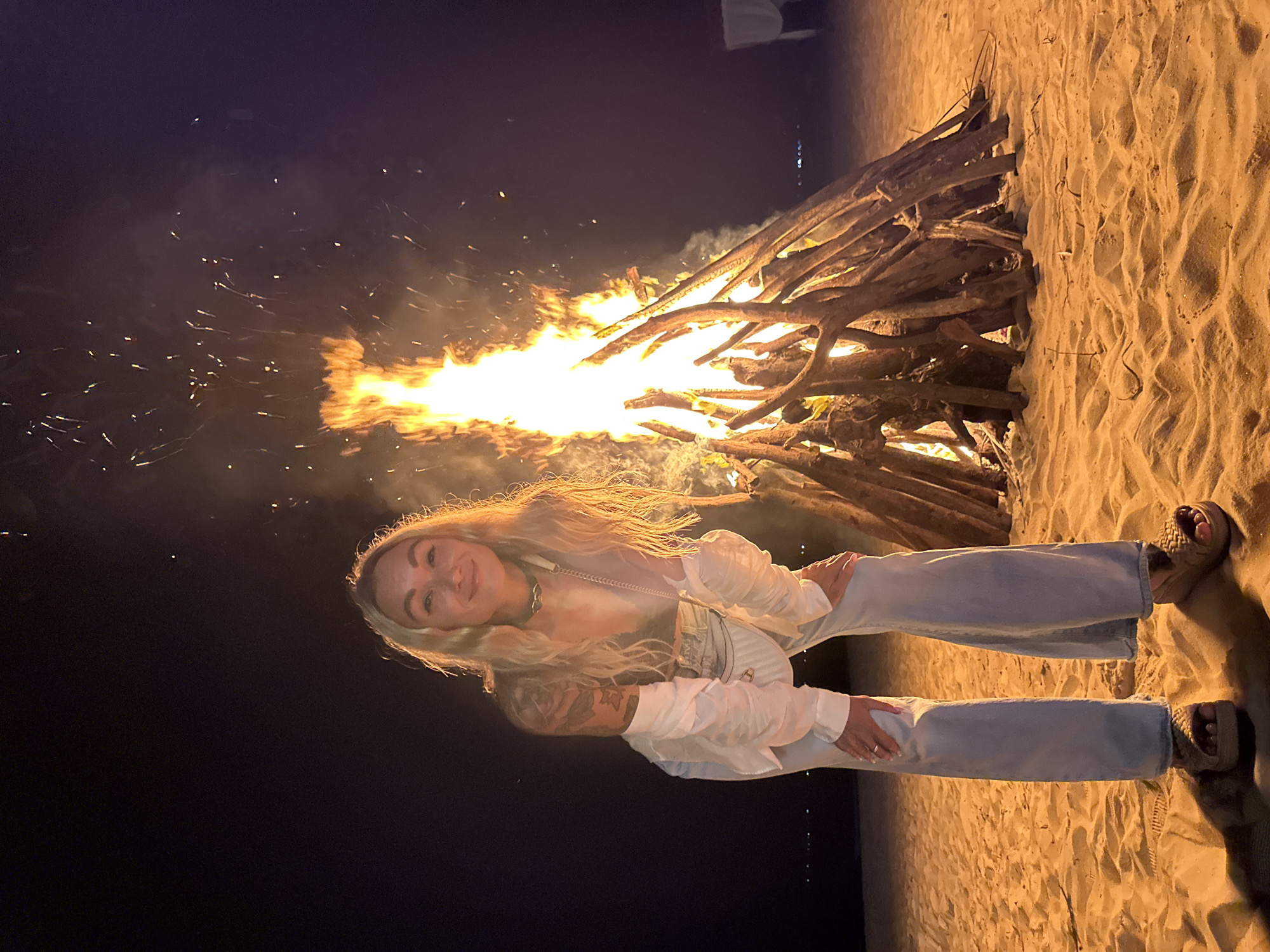 A woman with blonde hair and tattoos on her arms is posing in front of a large bonfire on a sandy beach at night. She is wearing a white shirt with puffed sleeves, light blue flared jeans, and platform sandals. Her hands are resting on her knees. The bonfire is made of large logs and is burning brightly, with flames and sparks rising into the dark sky. The beach appears to be in a coastal area, possibly Phu Thuy Ward, Vietnam, given the presence of sand and the ocean in the background, indicated by distant lights on the water. The overall scene suggests a nighttime gathering or event on a beach.