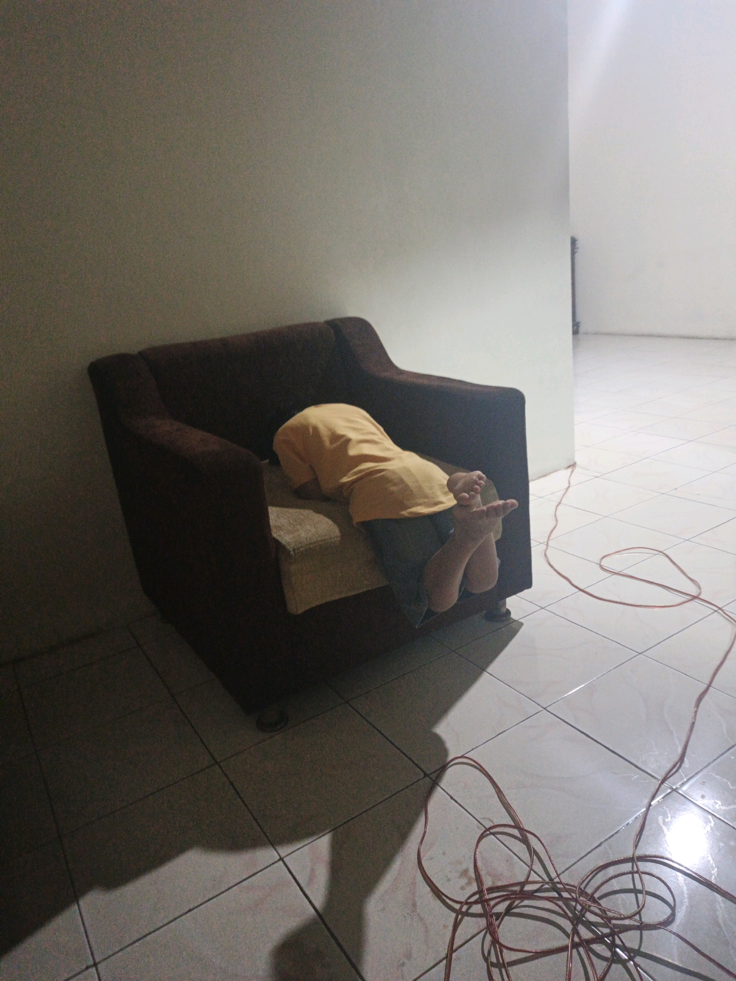In a sparsely furnished room with tiled flooring and light-colored walls, a single-seat armchair is positioned on the left side of the frame. A person, whose head is buried in the cushions of the armchair, is visible from the waist down. They are wearing a bright yellow shirt and dark pants. Their bare feet, with visible toes, are extended out from the side of the armchair, resting on the tiled floor. A tangled red electrical cord lies on the floor, stretching from the bottom right corner of the image and looping across the tiles. The lighting is dim, casting shadows on the floor. The room appears to be located in Bumiharjo, Indonesia, based on the provided context.