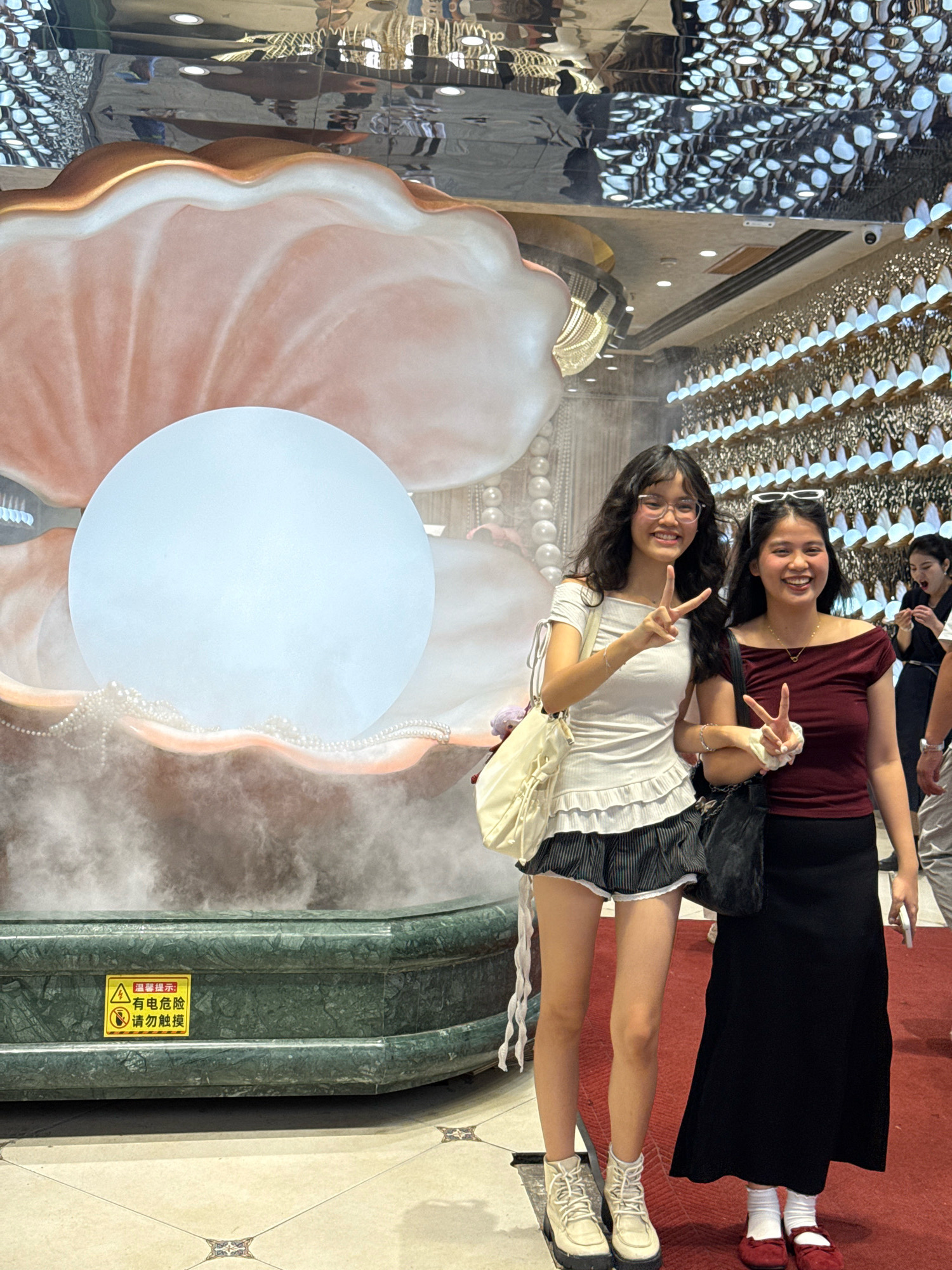 In Shanghai, China, two young women pose for a photo in front of a large, decorative oyster and pearl display. The woman on the left wears a white off-the-shoulder top with ruffled trim and a black and white striped skirt. She carries a cream-colored shoulder bag and wears glasses. The woman on the right is in a burgundy off-the-shoulder top and a long black skirt, holding a black handbag. Both women are smiling and making peace signs. The oyster display features a large, illuminated pearl surrounded by misty vapor and strings of faux pearls. A sign with Chinese characters, indicating a warning about electricity and advising against touching, is affixed to the green marble base of the display. The background shows a highly reflective ceiling and ornate wall decorations.