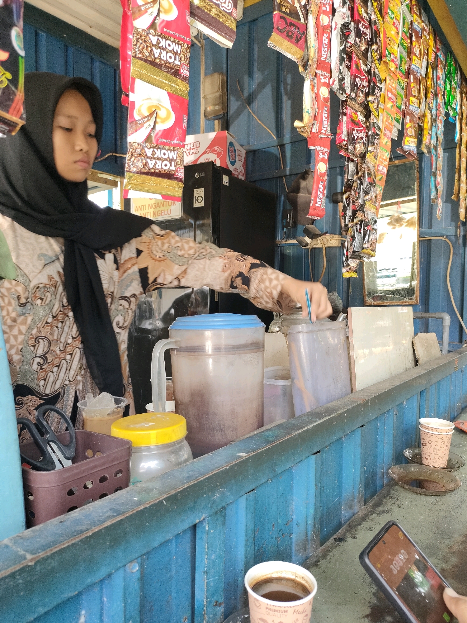 A young Indonesian woman wearing a black hijab and a patterned blouse stands behind a counter in Sukodono, Indonesia. She is holding a blue utensil and appears to be stirring a beverage in a large, clear plastic jug with a blue lid. In front of her, there are several items, including a brown basket containing a pair of black scissors, a clear plastic cup with ice and liquid, and a glass jar with a yellow lid. The counter is lined with various packaged food and drink items hanging from above, including sachets of coffee and snacks. Behind the woman, a black electronic device with an LG logo is visible. The counter itself is made of blue corrugated metal. On the far right of the counter, there are two cups of coffee and a small saucer. In the foreground, a smartphone displaying a timer is being held by an unseen hand. The overall environment suggests a small roadside stall or convenience store.