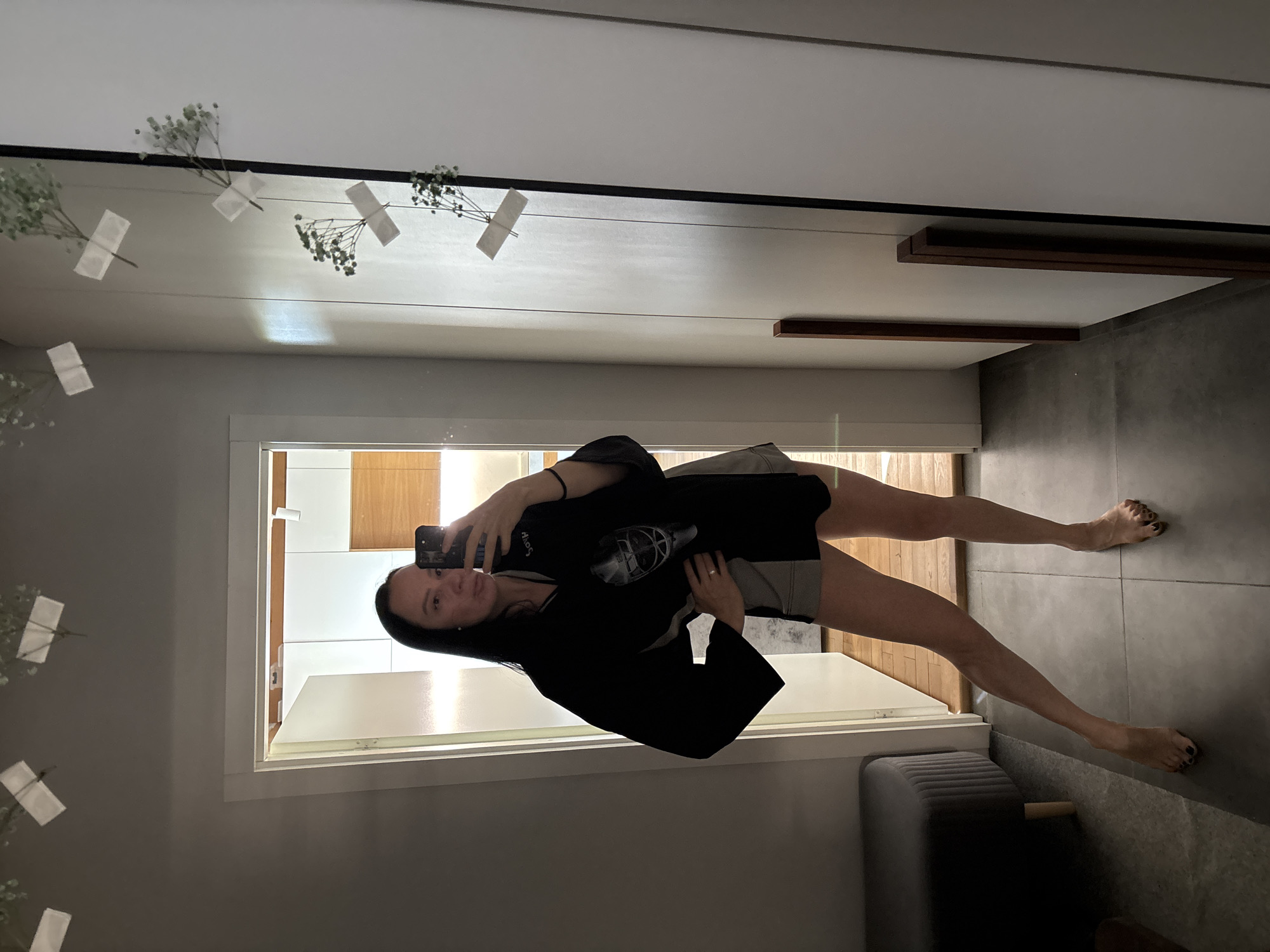 A medium shot depicts a woman taking a selfie in front of a mirror. The mirror is adorned with decorations, including small white flowers taped to the top. The woman is wearing a black top over shorts. She is holding a smartphone in her right hand, using it to capture the image. Behind her, visible through a doorway reflected in the mirror, is a room with wood-paneled walls and white fixtures. A gray upholstered ottoman is visible in the foreground. The location is identified as the city of Chaikovsky, Russia.