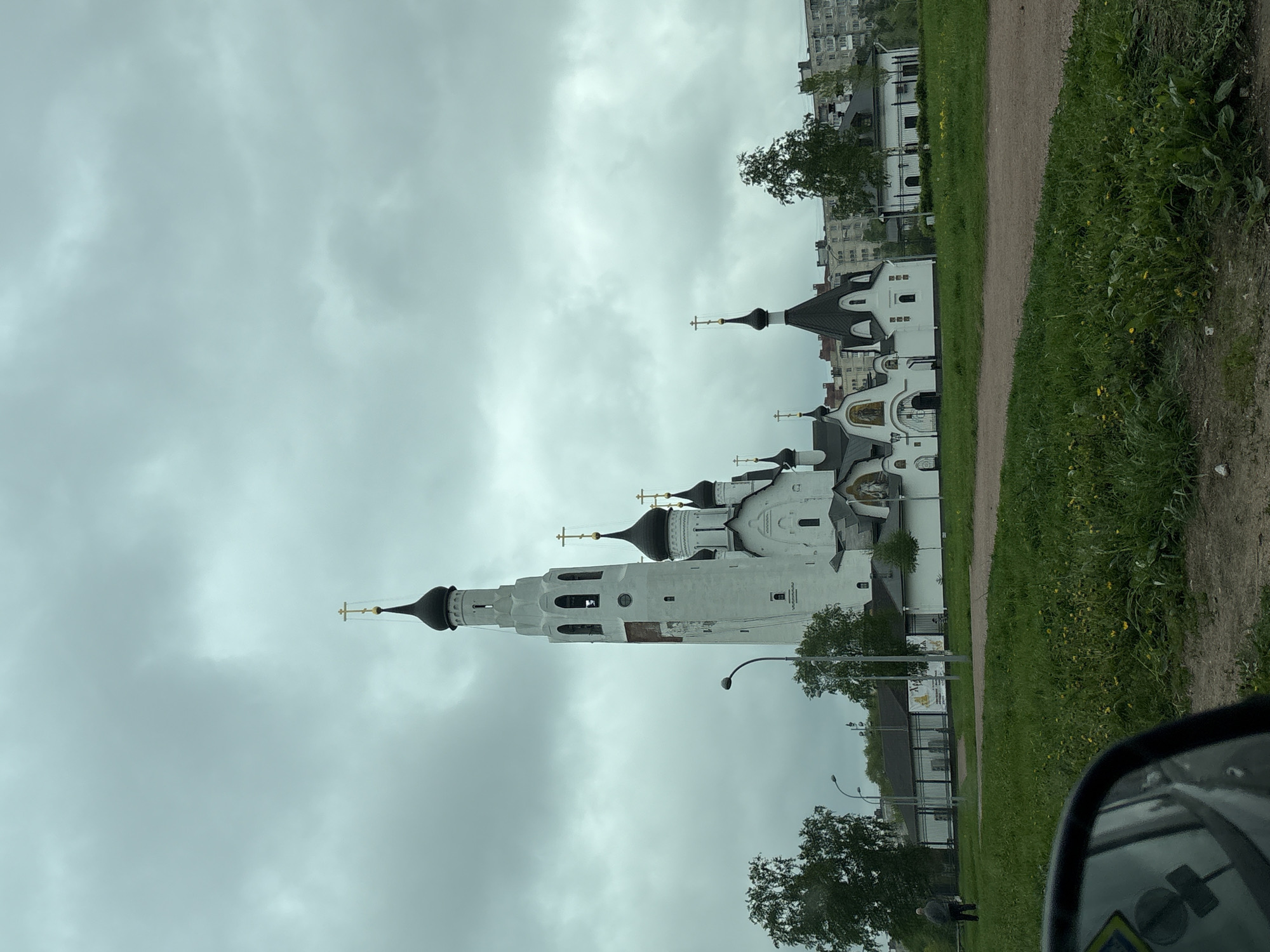 The image shows a tall, white Orthodox church complex under an overcast sky. The church features a prominent bell tower, black onion domes topped with gold crosses, and detailed architectural elements. The foreground includes a grassy area, a dirt path, and a glimpse of a car's side mirror. In the background, there are other buildings, possibly residential apartments. The location appears to be Saint Petersburg, Russia, based on the architectural style of the church and the overall environment.