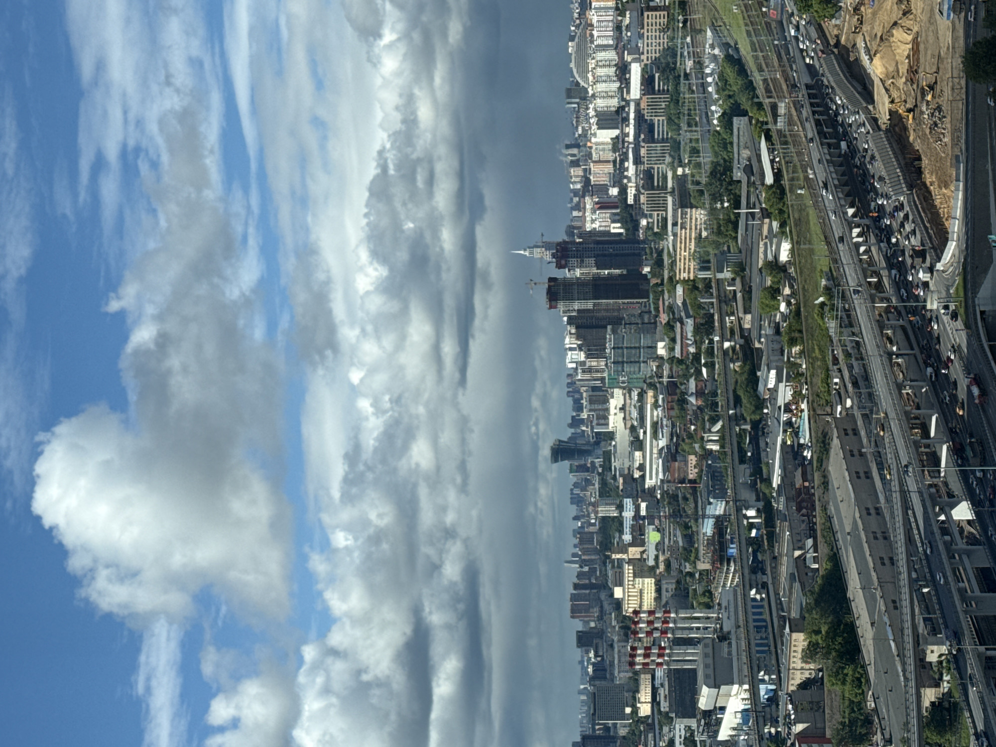The image shows an aerial view of a city with a dramatic sky overhead. The sky transitions from blue with white, fluffy clouds to a band of dark, stormy clouds. The city consists of numerous buildings, ranging in height and style. Roads and railway lines are visible, along with dense patches of greenery. There is a construction site on the right side of the image, which features dirt and heavy machinery. The scene does not take place in Khimki, Russia.