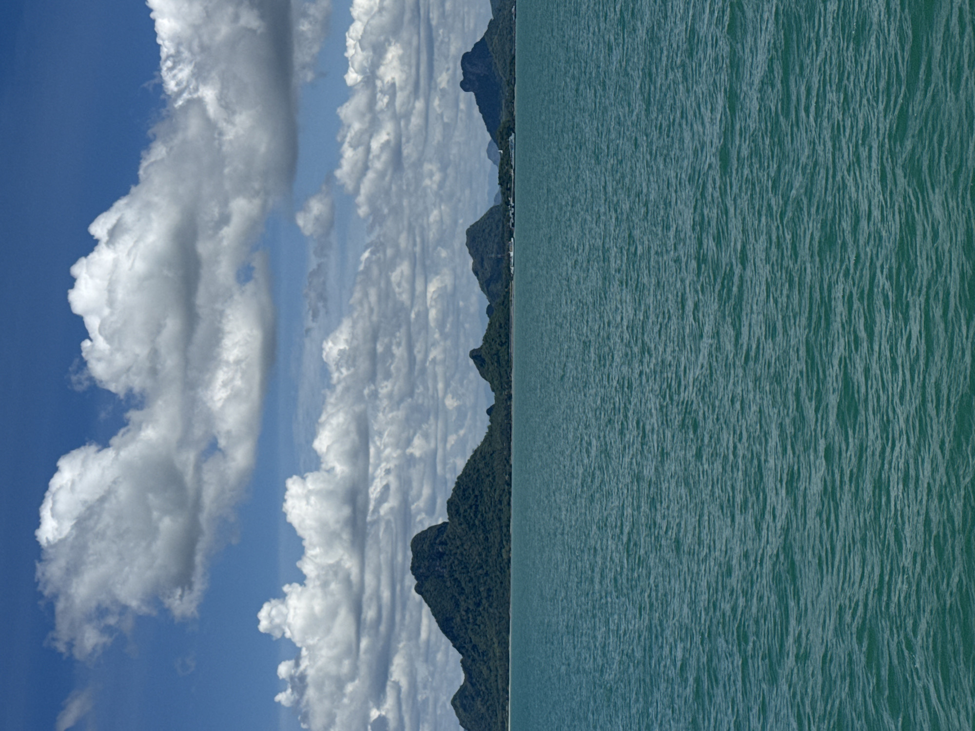 A vertical, medium shot captures a vast expanse of deep turquoise water in the foreground, with gentle ripples across its surface. In the middle ground, a lush, green mountainous island chain stretches across the horizon, dotted with trees and featuring distinct, rugged peaks. A narrow strip of sandy beach is visible at the base of the islands. Distant structures, possibly buildings or boats, are faintly discernible along the coastline. The background is dominated by a bright blue sky filled with large, fluffy white clouds, creating a dynamic contrast with the serene water and verdant islands. The scene appears to be a natural landscape, likely a coastal or island environment.