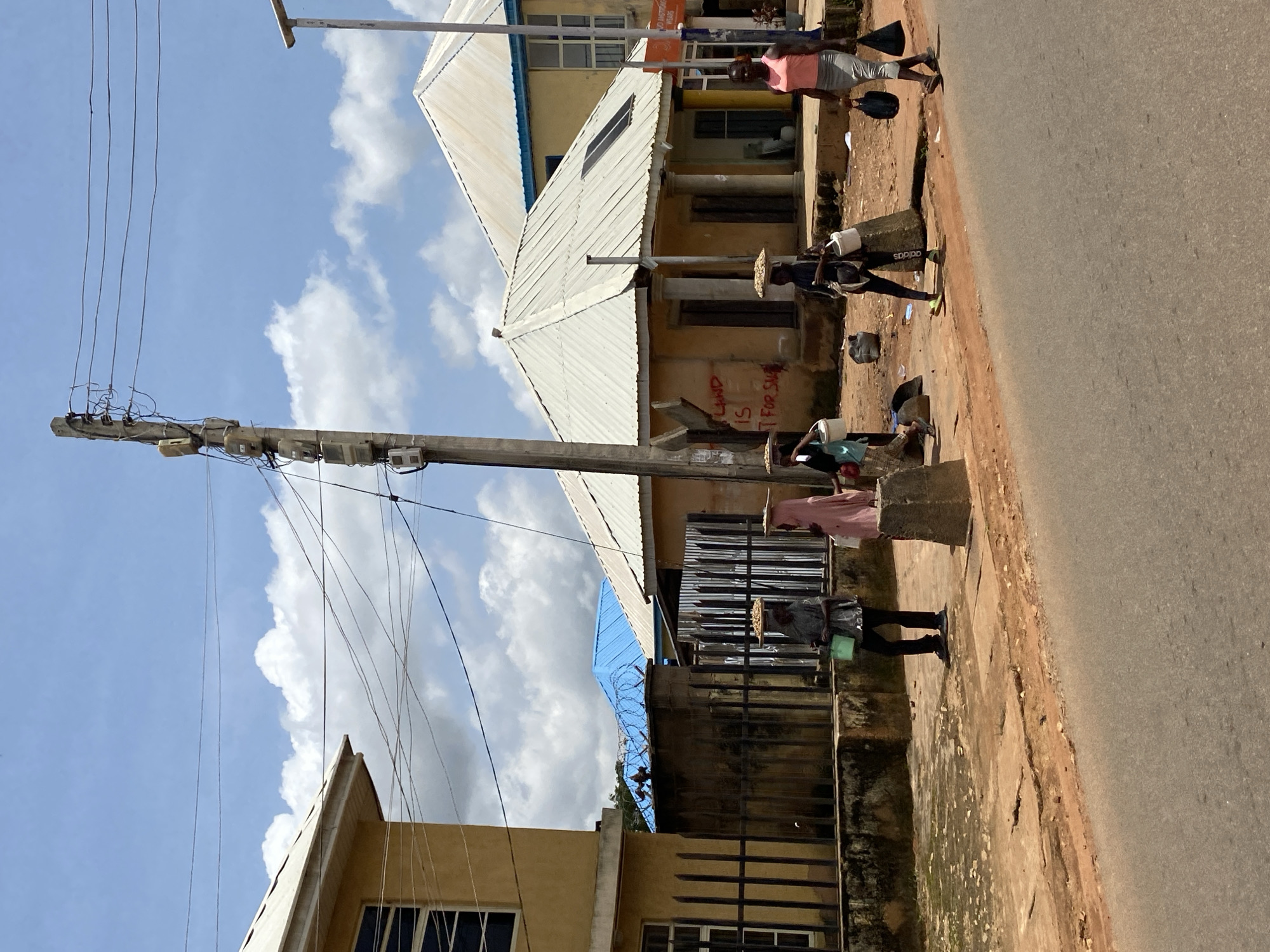 A medium-shot captures a street scene in Gura Top, Nigeria under a partly cloudy sky. In the foreground, a paved road runs diagonally across the frame from bottom left to top right. The middle ground features a raised dirt sidewalk with scattered debris and vegetation. A line of buildings runs along the sidewalk. The building on the far left is yellow with a dark-framed window. Adjacent to it is a fence and a wall topped with blue barbed wire. Further down the street are structures with white corrugated metal roofs. One building has "LAND IS FOR SALE" scrawled on its exterior wall. A utility pole with multiple meters and tangled wires stands prominently on the sidewalk.
Four individuals are visible walking on the sidewalk. The first person from the left, dressed in a grey shirt and dark pants, carries a green bucket and a bowl on their head. Next, a person in a pink veil and dress also carries a bowl on their head. Following them, a person in black athletic wear with "adidas" visible on their leg carries a bowl on their head and a white bucket. The person furthest to the right wears a pink tank top and a grey skirt, carrying two black bags. Bags and other objects are also scattered on the ground near the people. The overall impression is a typical day on a street in a Nigerian city, with individuals engaged in daily activities.