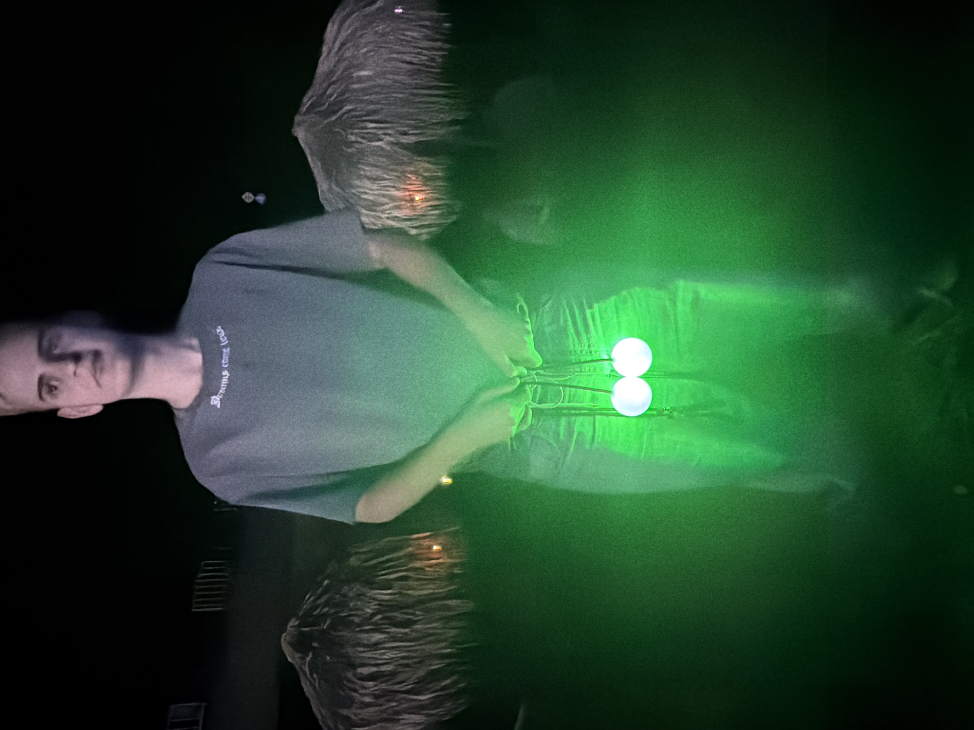 A person stands in front of two tiki umbrellas at night, holding two glowing white balls on strings. The person is wearing a grey t-shirt with white text that says "Dreams come true" and blue jeans. The scene is illuminated by green light, likely from the balls themselves or ambient lighting. The background is dark and indistinct, with some faint structures visible behind the tiki umbrellas, potentially part of a resort or outdoor venue in Zgorigrad, Bulgaria.