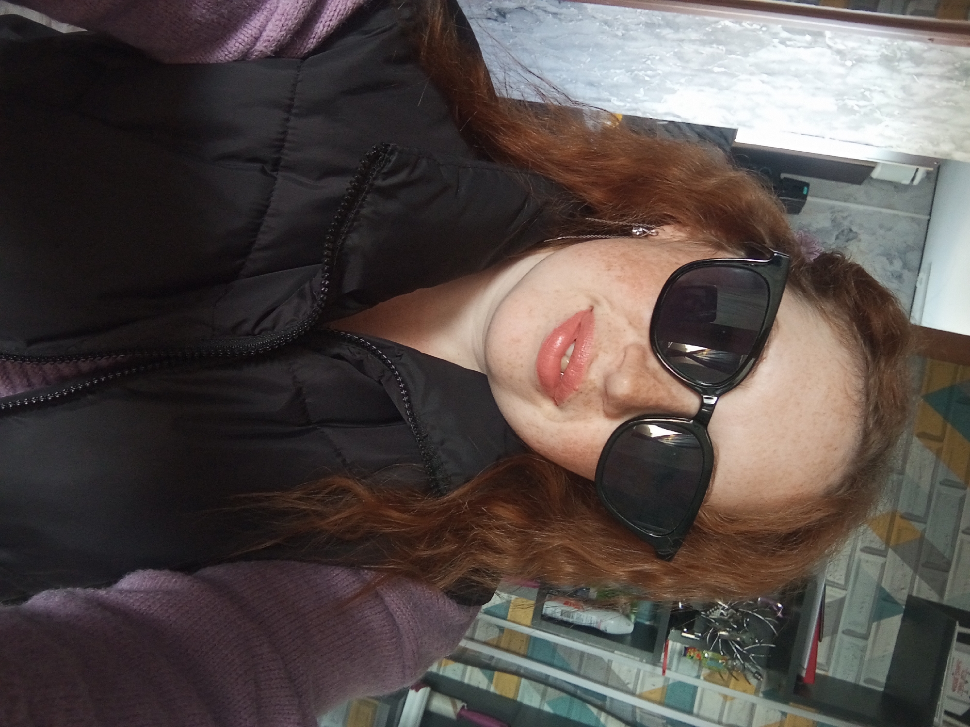 A red-haired woman with freckles is wearing sunglasses and a black puffy vest over a light purple sweater, posing for a selfie in a bathroom, likely located in Yurgamysh, Russia. Behind her is a wall covered in geometric patterned wallpaper and a vanity area containing items on display.