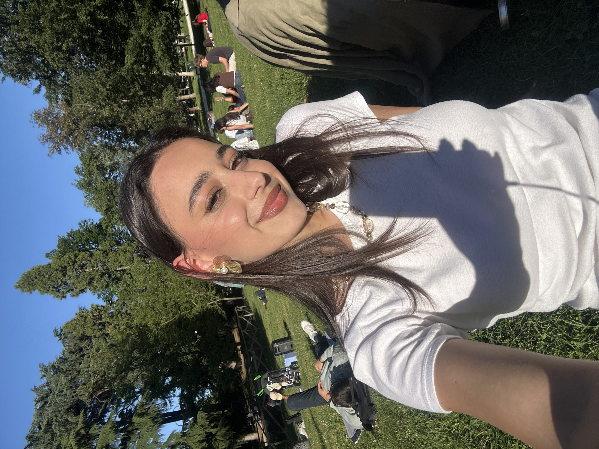 A selfie shows a young woman with long, dark hair wearing a white t-shirt and gold floral earrings, smiling. She is lying on a grassy area in what appears to be a park. In the background, other people are also relaxing on the grass. Trees are visible in the background. The photo was taken in Milan, Italy.
