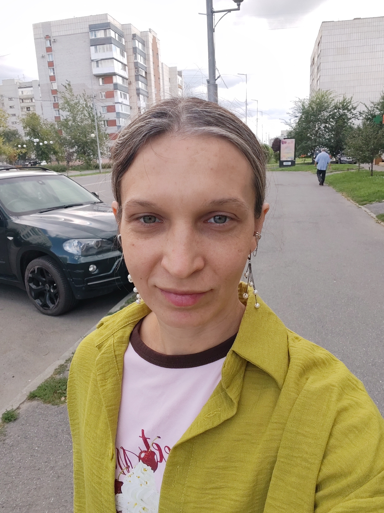 A medium shot photograph captures a woman with blonde hair, wearing a green jacket and a pink t-shirt with cherries, standing on a sidewalk next to a dark green SUV. She is looking directly at the camera, with her left ear adorned with pearl earrings. In the background, a man in a blue shirt is walking away from the camera on the sidewalk, and several apartment buildings and trees are visible. The sky is overcast.